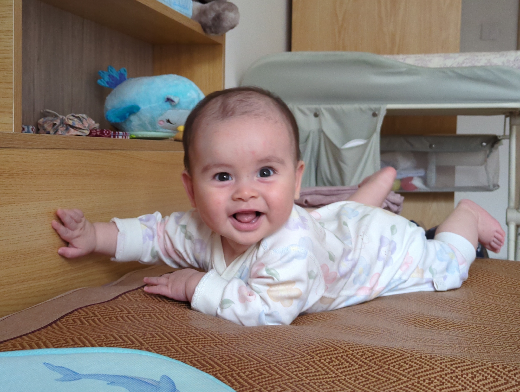 A baby lies prone on a brown patterned surface. The baby wears a white outfit patterned with light-colored floral designs. The baby's head is raised, with an open mouth and a visible smile. The baby's left hand touches the side of a wooden structure, while the right hand rests on the surface. A blue stuffed animal with a shark design is partially visible in the lower left corner of the frame. In the background are wooden shelves with stuffed animals and a changing table. The location cannot be forensically verified as Shunde District, China.