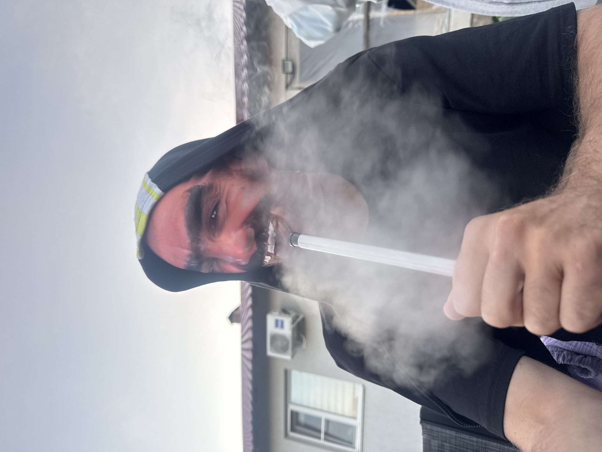 A man, with his face painted red and wearing a dark hooded shirt, is using a white smoking device, which is producing a plume of smoke. He is outdoors, in front of a building with a tiled roof and an air conditioning unit. The sky is overcast. The location is identified as Узюково, Russia.