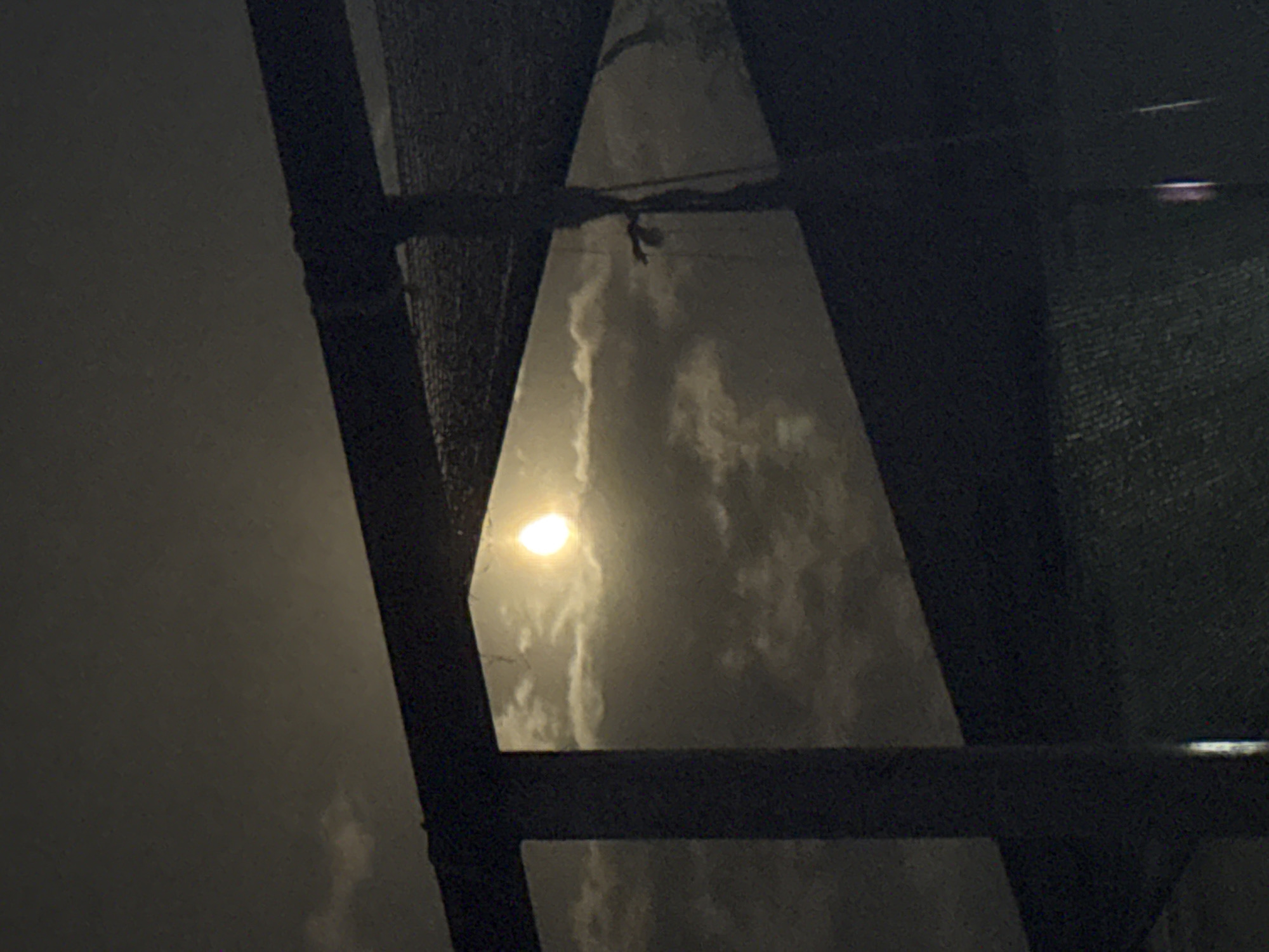 The image depicts a nighttime scene viewed through a dark, rectangular frame, possibly a window or an opening in a structure. The sky is visible, illuminated by a bright, round object which appears to be the moon, partially obscured by clouds. The clouds themselves are wispy and illuminated by the moon's light. The foreground is dominated by dark, shadowy structures, suggesting an urban or semi-urban environment. The overall impression is one of a serene, nocturnal landscape, with the moon as the focal point. There is no indication of the specific location being Vostok, Kyrgyzstan.