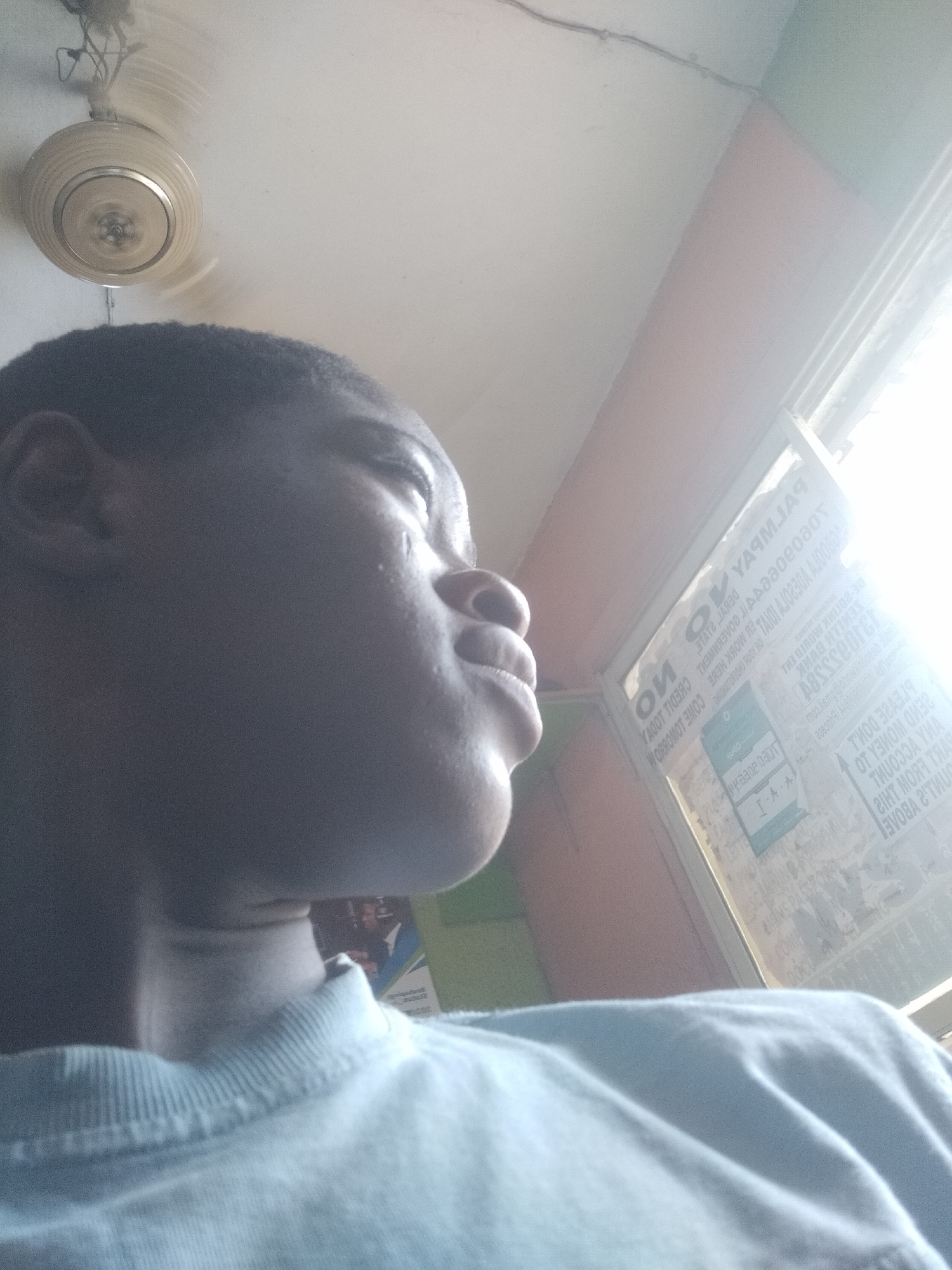 A low-angle, close-up shot of a Black person's face, looking upward. A ceiling fan is visible in the upper left corner. A window with printed signs is to the right, with illegible text due to overexposure. The person is wearing a light blue t-shirt. The location is assumed to be in Shangisha, Nigeria, based on the textual elements on the glass.
