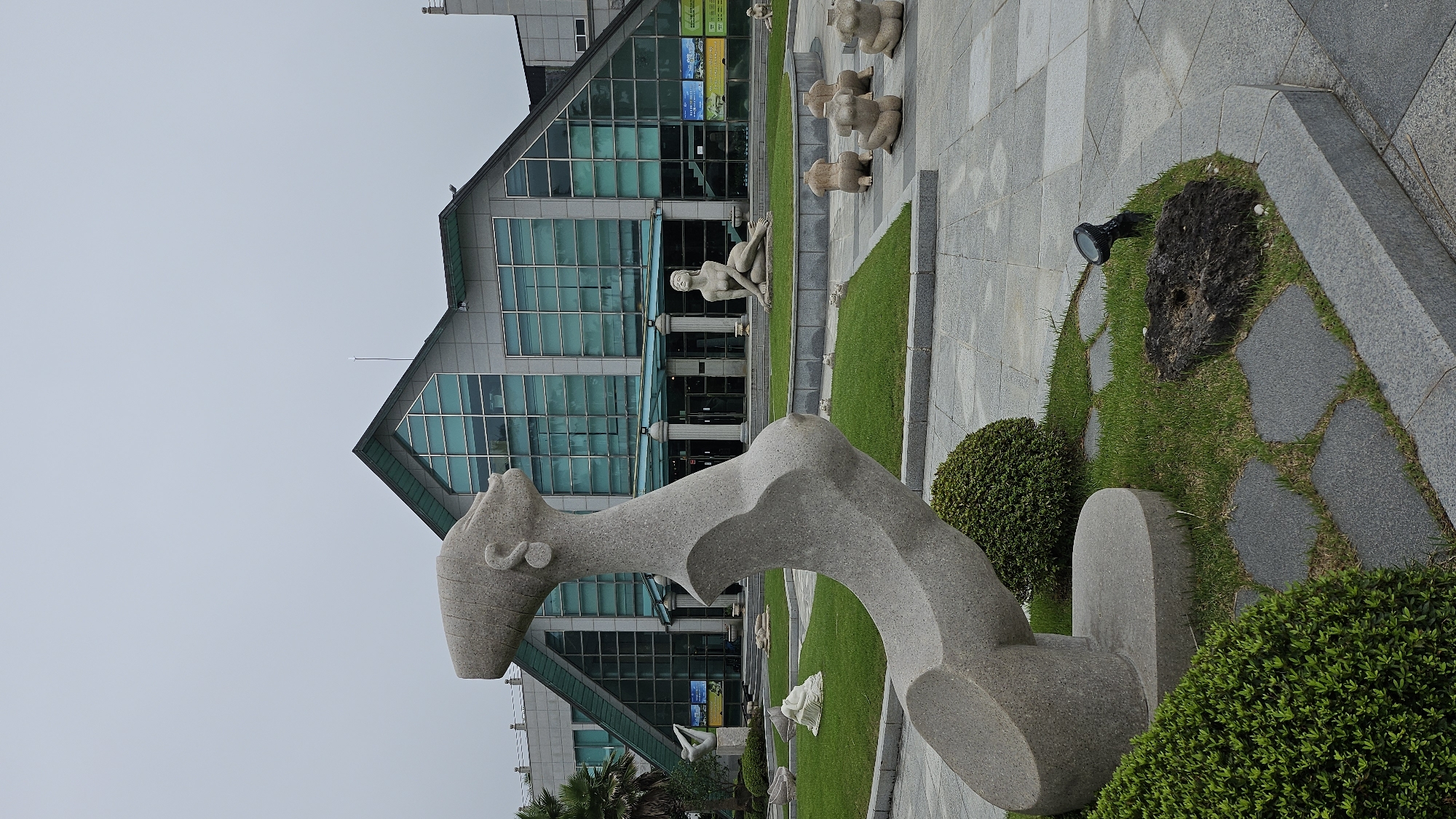 The image depicts the exterior of the Seogwipo Arts Center, located in Seogwipo-si, South Korea. The building features a modern architectural design with a large, angled glass facade. In the foreground, a large, abstract stone sculpture resembling a stylized human figure is prominently displayed. Several other stone sculptures, primarily of seated female figures, are arranged on the surrounding lawn and paved areas. The environment is characterized by neatly manicured grass, paved walkways, and some greenery. The sky is overcast, suggesting a cloudy day. No people are visible in the image.