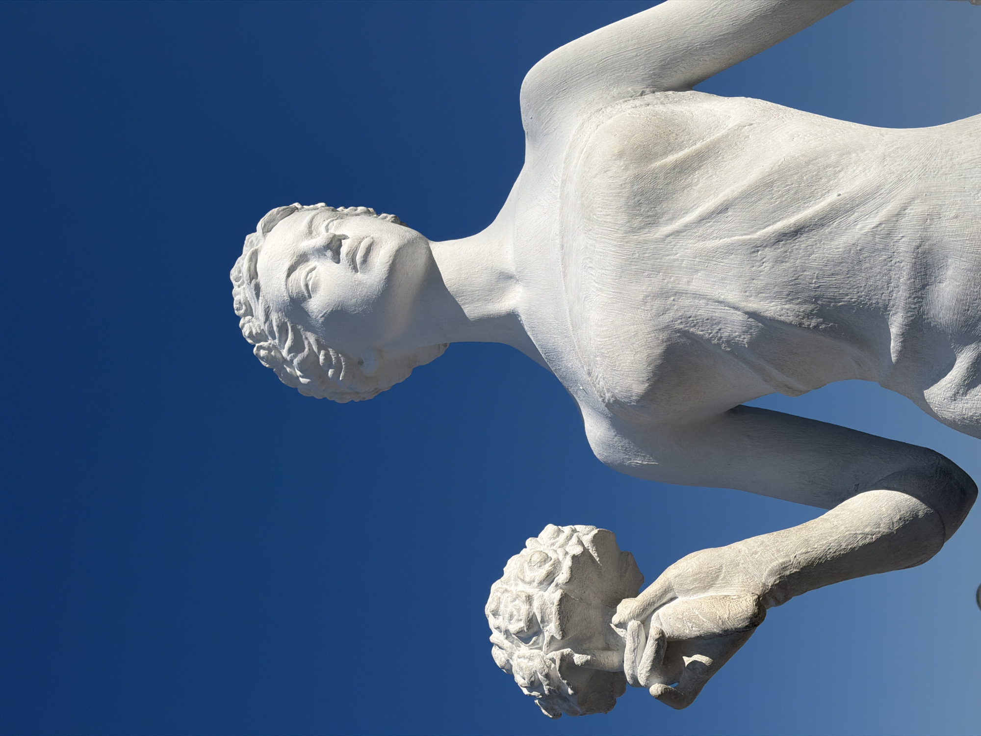 This is a low-angle, medium shot of a white stone statue of a woman against a clear, vibrant blue sky. The statue is positioned on the right side of the frame, with its head tilted slightly upwards and to the left. The woman has curly hair and is holding a bouquet of flowers in her left hand, which is positioned in the lower left quadrant of the image. The statue has a textured surface, with visible chisel marks, particularly on the skin and the flowers. The sky is a solid, deep blue, with no clouds or other atmospheric phenomena. The statue is situated outdoors, suggesting it is a public sculpture, likely in Gelendzhik, Russia, a city known for its numerous sculptures and seaside promenade. The lighting indicates a bright, sunny day.