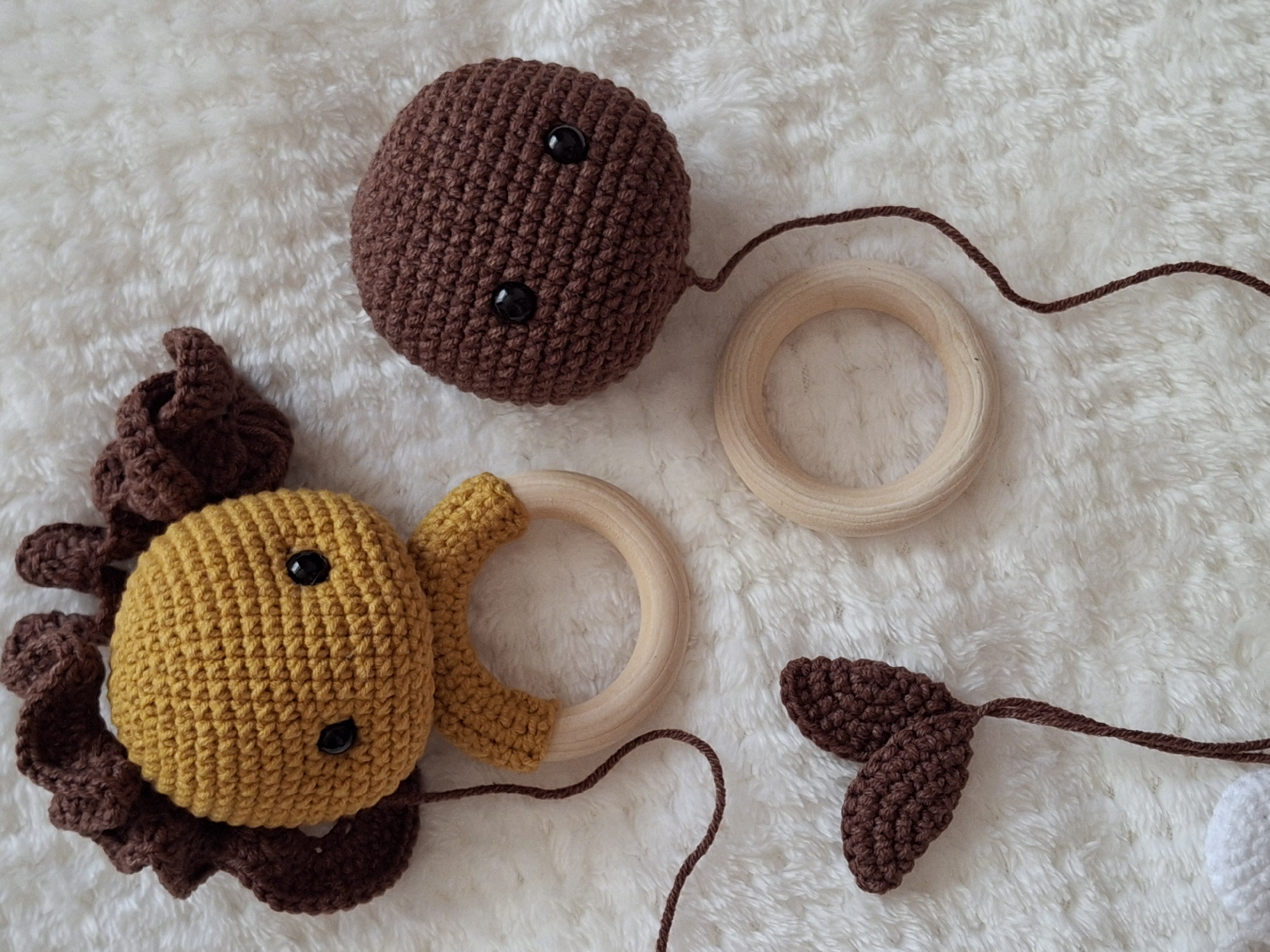 The image shows crocheted baby toys, possibly a mobile, arranged on a white, fluffy surface. The toys include a brown sphere with two black button eyes, a yellow sphere with a brown mane and two black button eyes, two wooden rings, and brown crocheted leaves. Brown string connects the elements. The location, Chaikovsky, Russia, is unrelated to the image content.