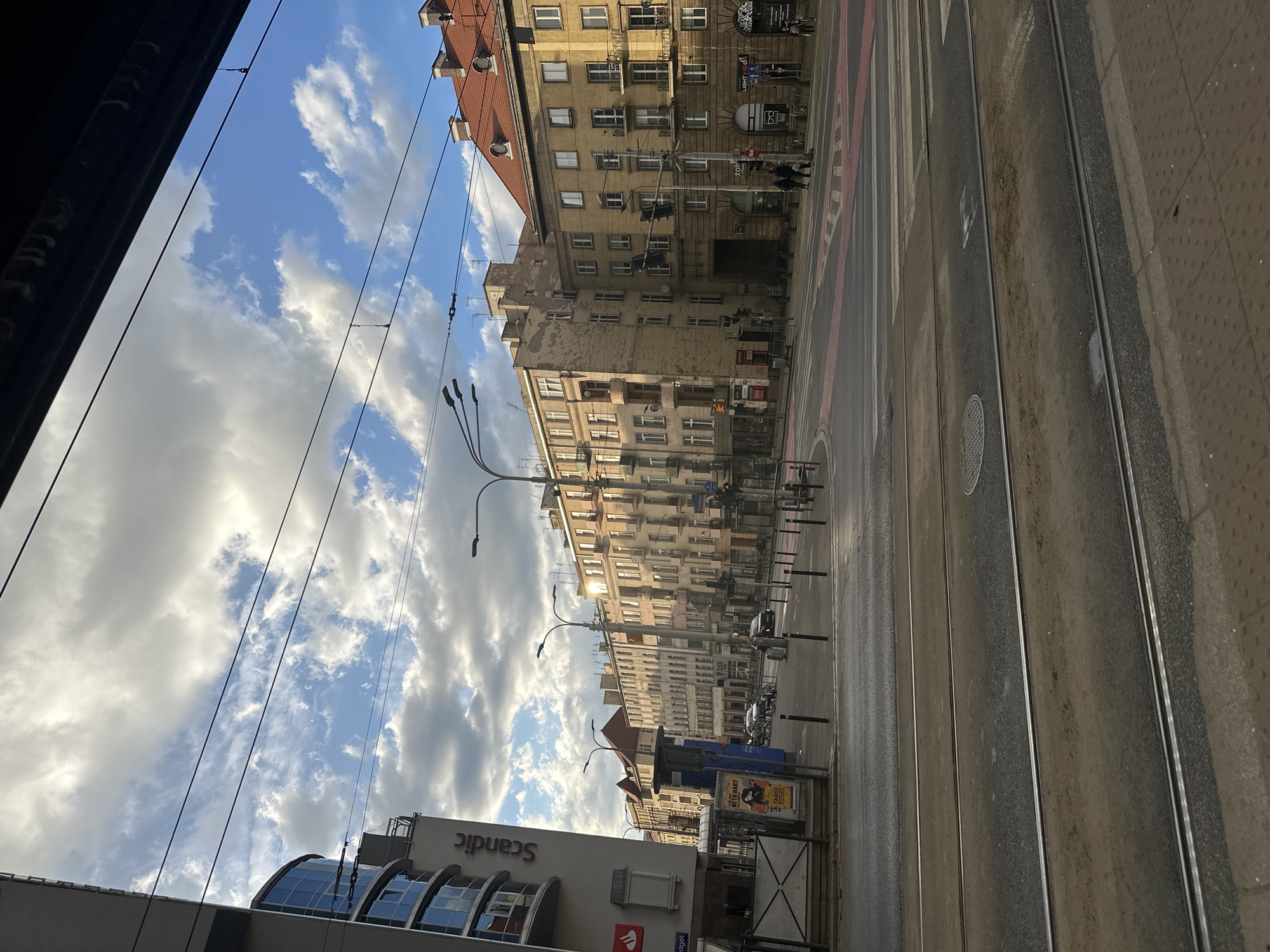The image depicts a street scene in Wrocław, Poland. The view is taken from an elevated position, possibly inside a tram or building. On the right is a street with tram tracks running along it. Adjacent to the street is a sidewalk made of concrete pavers. Along the left are buildings of varying heights and architectural styles. In the background, there are older, multistory buildings with multiple windows and balconies. Closer to the viewpoint, there is a more modern building with a glass facade and the "Scandic" brand prominently displayed. Utility poles with overhead wires are visible, traversing the sky filled with clouds. A few people are walking along the street.