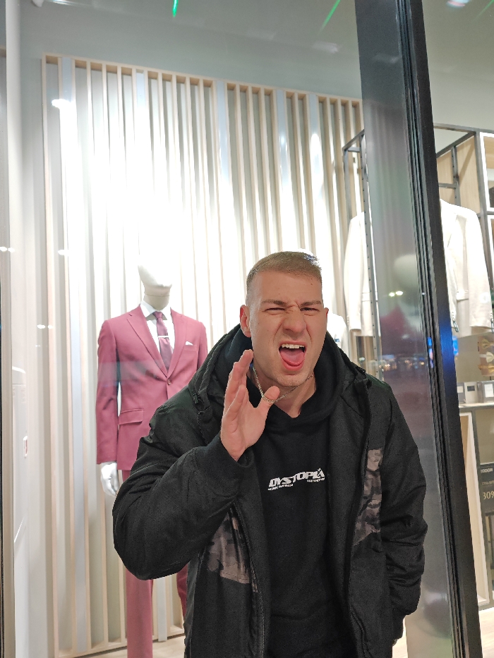 An individual, identified as a young adult male with short, light-colored hair, stands directly in front of a retail storefront or display window. He is wearing a dark hooded sweatshirt with white text "DYSTOPIA" and a dark puffer jacket, accessorized with a silver chain around his neck. His facial expression is exaggerated, with eyes squeezed shut and tongue extended. His right hand is raised to chest height, palm open and fingers splayed.
Behind him, through the clear glass of the storefront, a retail interior is visible. To the left, a male mannequin is dressed in a maroon suit, white shirt, and dark tie with a tie clip, positioned against a backdrop of vertical light-colored slats. To the right, beyond a dark vertical frame, additional light-colored garments on racks are discernible. Reflections on the glass surface show faint green light streaks, suggesting artificial lighting within an indoor commercial setting, likely a shopping mall. The image contains no explicit visual markers to definitively pinpoint its location within Shumen, Bulgaria.