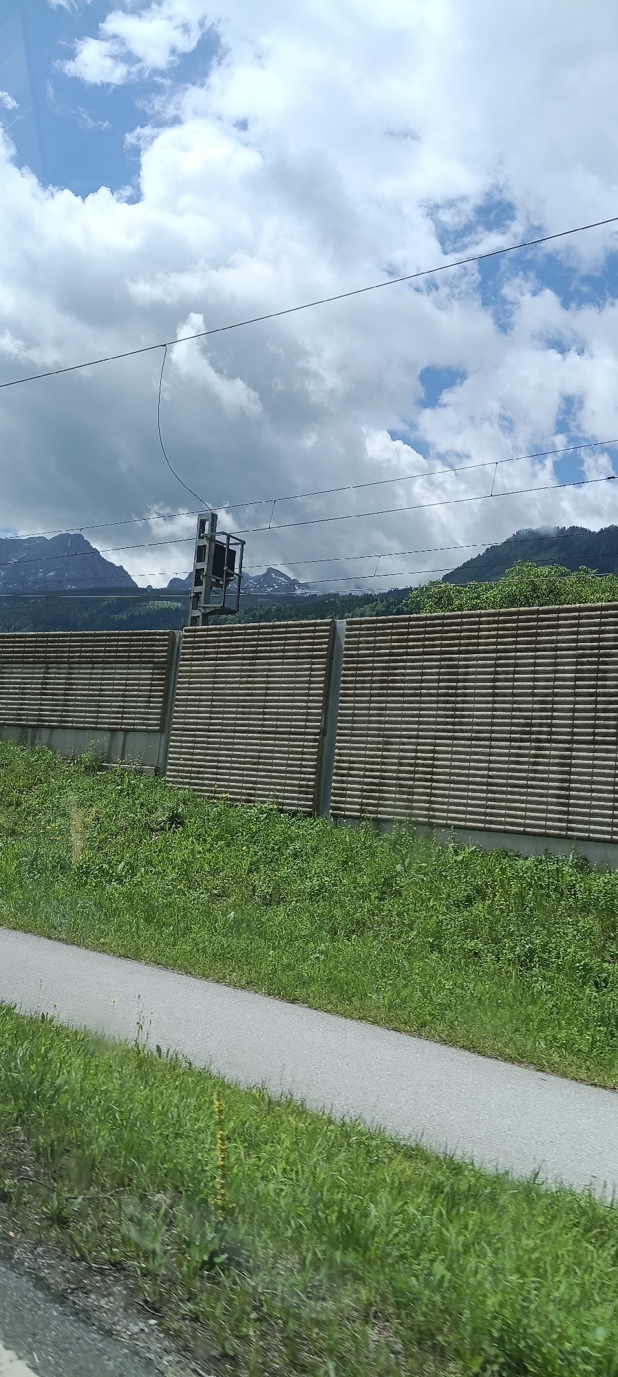 The image shows a view from a roadside in Moos-Siedlung, Austria. In the foreground, there is a paved path running diagonally from the bottom left to the top right, bordered by green grass. Behind the path and grass is a tall, segmented sound barrier constructed of horizontal wooden slats. Beyond the sound barrier, a dense line of green trees and foliage is visible, with the lower slopes of mountains in the background. The mountains are covered in patches of snow at their higher elevations, and the sky above is a mixture of blue and dramatic, dark clouds, with some white, puffy clouds. Overhead, power lines for a railway are suspended from poles. A signal pole with a black signal head is also visible above the sound barrier, indicating railway infrastructure.