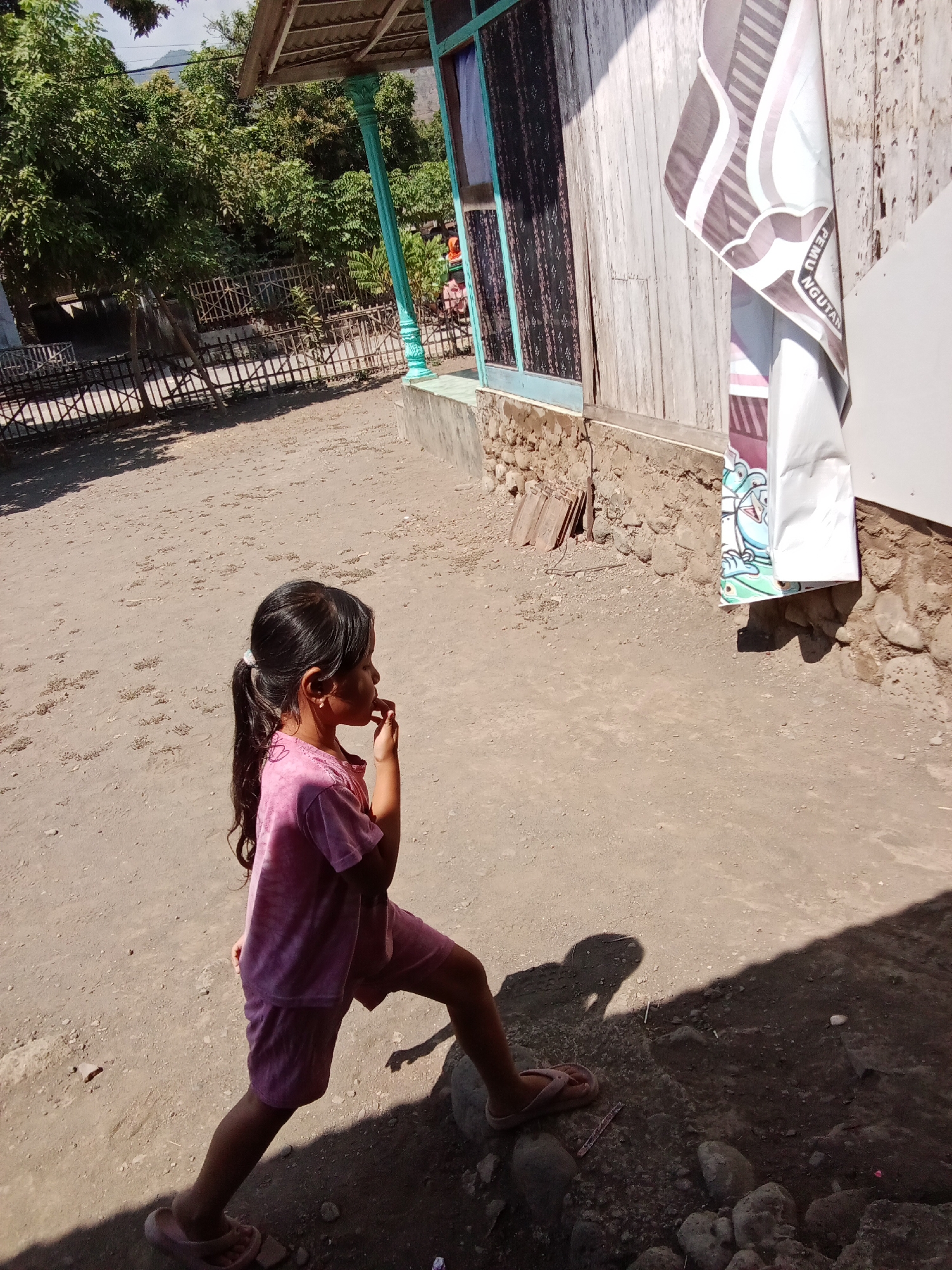 In Trebungan, Indonesia, a young girl with dark hair in a ponytail, wearing a pink t-shirt and shorts, stands in a dirt yard. She is facing right, with her right hand touching her mouth. Her left leg is extended forward. A shadow of the girl is cast on the ground to her right. Behind her, to the left, is a wooden fence and lush green trees. To the right of the girl, a rustic wooden building stands with a tiled roof. A banner is hanging on the side of the building, partially obscuring a window with a dark patterned curtain. The building's foundation is made of stone. The ground is composed of dry dirt with scattered small rocks.