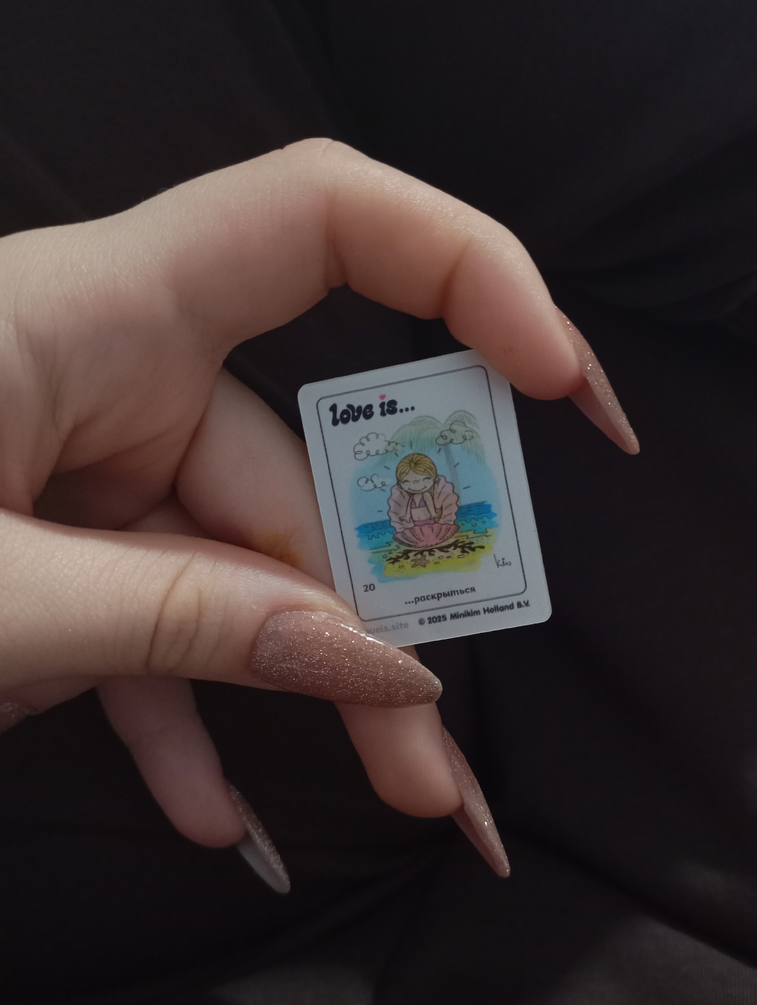 A fair-skinned hand with long, glittery, brown-ombre nails holds a small white card. The card features an illustration with the text "Love is..." at the top. The illustration depicts a blonde-haired girl sitting on a sandy beach with her arms crossed, sheltering inside a shell. There are stylized waves and clouds behind her. The number "20" is printed in the lower-left corner, and below the illustration, there is Russian text and the copyright information "© 2023 Minikim Holland B.V." The background is dark and out of focus. There's no indication of Krasnodar, Russia.