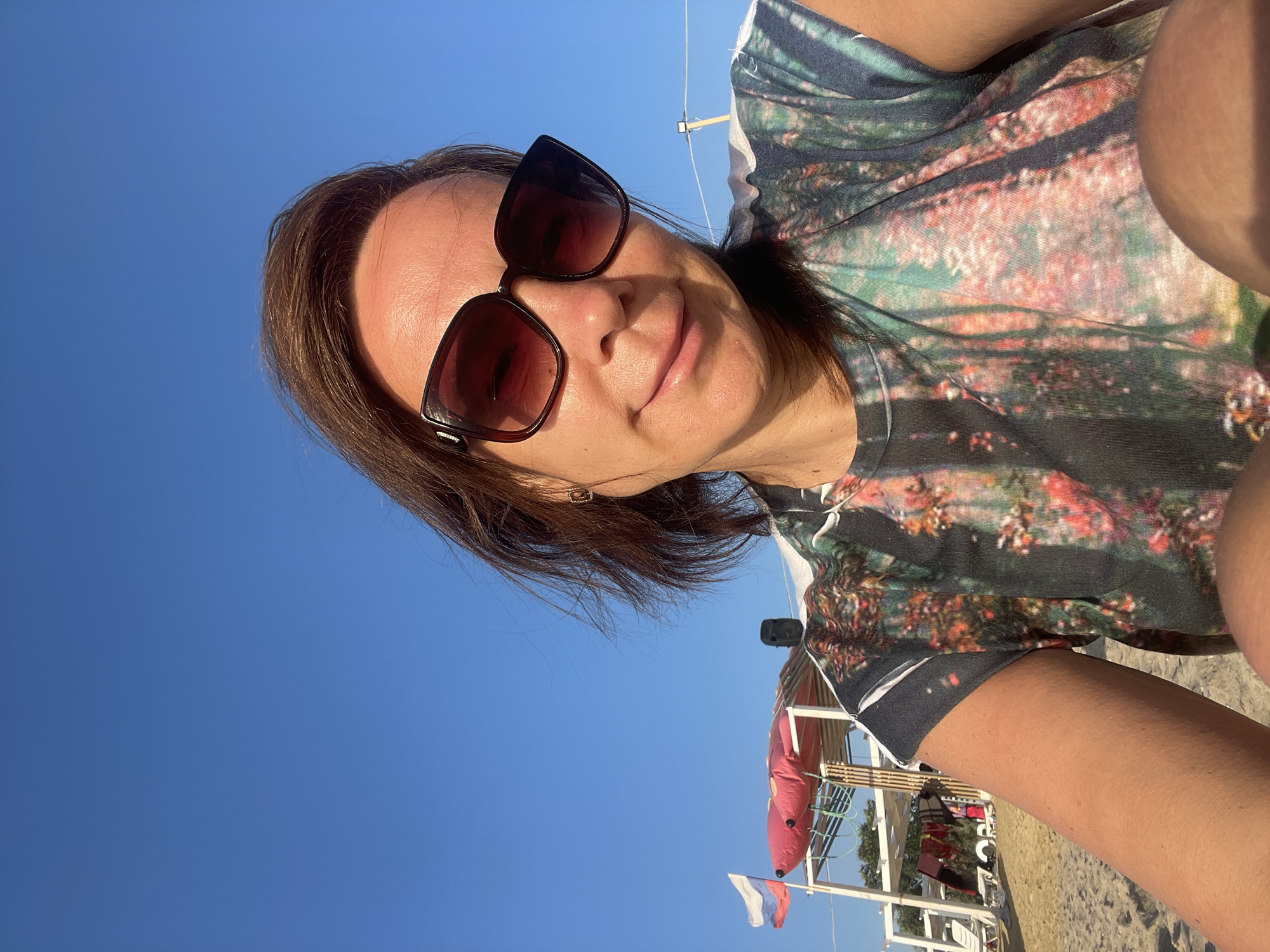 A medium shot captures a woman with medium-length brown hair, wearing large, dark-rimmed sunglasses and a t-shirt with a colorful floral print. She is outdoors, seated on a sandy beach with a bright blue sky overhead. In the background, a white structure with a red canopy is visible, along with beach chairs and the Russian flag flying on a pole. The woman is looking directly at the camera, with a slight smile on her face. The scene suggests a leisurely day at the beach in Anapa, Russia.