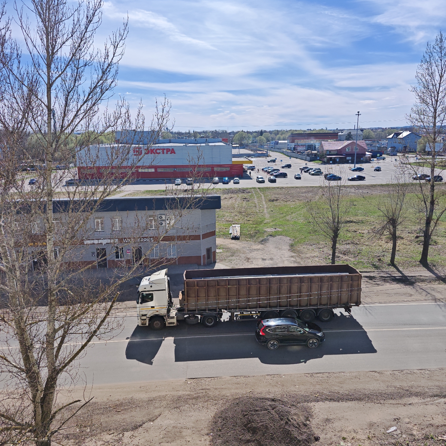 This elevated view captures a bustling commercial and light industrial area in Rostov, Russia, under a bright, partly cloudy sky. The scene depicts a typical daytime setting, likely late morning or early afternoon, evidenced by the distinct shadows cast by objects and vehicles.
In the foreground, the bare branches of a tree frame the left side of the image, suggesting either early spring before leaf-out or late autumn. A two-lane road stretches across the midground, where a white semi-truck with a large, dark brown, open-top trailer is seen driving. Alongside it, a dark-colored SUV, possibly a Honda CR-V, also travels down the road, with both vehicles casting long shadows.
Immediately adjacent to the road on the left, a two-story building with a grey facade displays the English text "WILD BERRIES," indicating a facility for the Russian e-commerce giant.
Further into the background, a large commercial building dominates the landscape with a prominent red and white sign featuring the Cyrillic text "ЭКСТРА" (EKSTRA), suggesting a large store or supermarket. Multiple parking lots surround this building and other smaller commercial structures, populated with numerous parked cars. Beyond these immediate buildings, a mix of further urban development, sparse trees, and grassy patches extends to the horizon.
The image portrays a routine day in a commercial zone, with vehicles on the road and parked in lots, indicating ongoing daily activities. The weather is fair and bright, illuminating the various structures and natural elements within the frame.