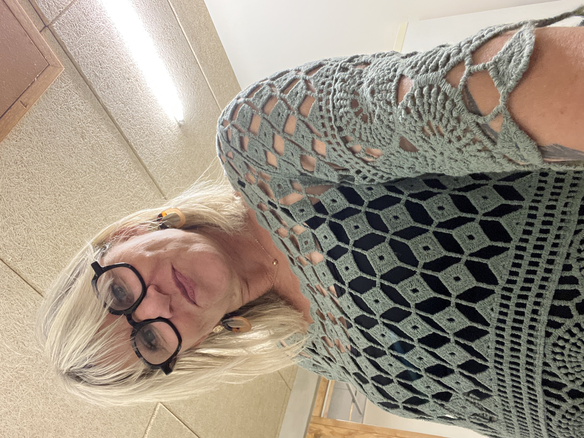 An upward-facing, low-angle shot captures a person with fair skin and short, layered blonde hair, wearing dark-rimmed glasses. The individual is looking directly towards the camera with a neutral expression, suggesting a slight smile. They are wearing a light green or sage green crocheted garment, possibly a top or sweater, which features an intricate openwork pattern of interconnected diamond shapes. This pattern reveals a dark garment worn underneath. Small, decorative earrings are visible on both ears.
The background consists of a textured, light-colored acoustic ceiling, typical of commercial or institutional buildings. A long, bright fluorescent light fixture is prominently visible across the ceiling. A wooden frame or panel is partially seen in the upper left corner of the frame. The lighting is artificial and bright. The scene is an indoor setting, with no specific activity other than posing for the photograph. No text is visible in the image. The location context provided is La Fouillouse, France.