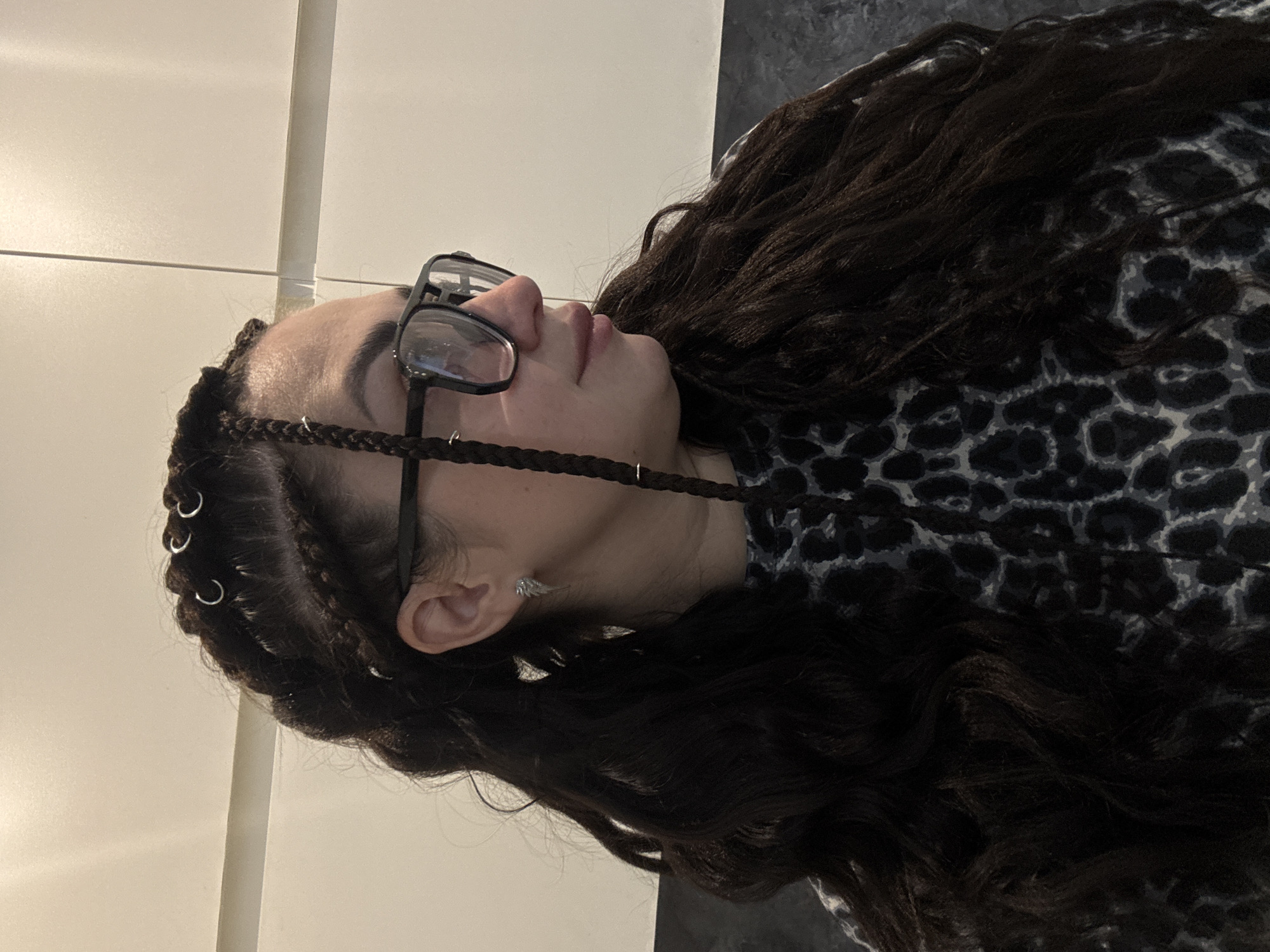 A woman with dark, wavy hair is pictured in a close-up, profile view. She is wearing glasses with thick black frames and a black and grey leopard-print top. Her hair is styled in braids, some of which are adorned with small metal rings. She has a silver earring in the visible ear. The background appears to be an interior wall.