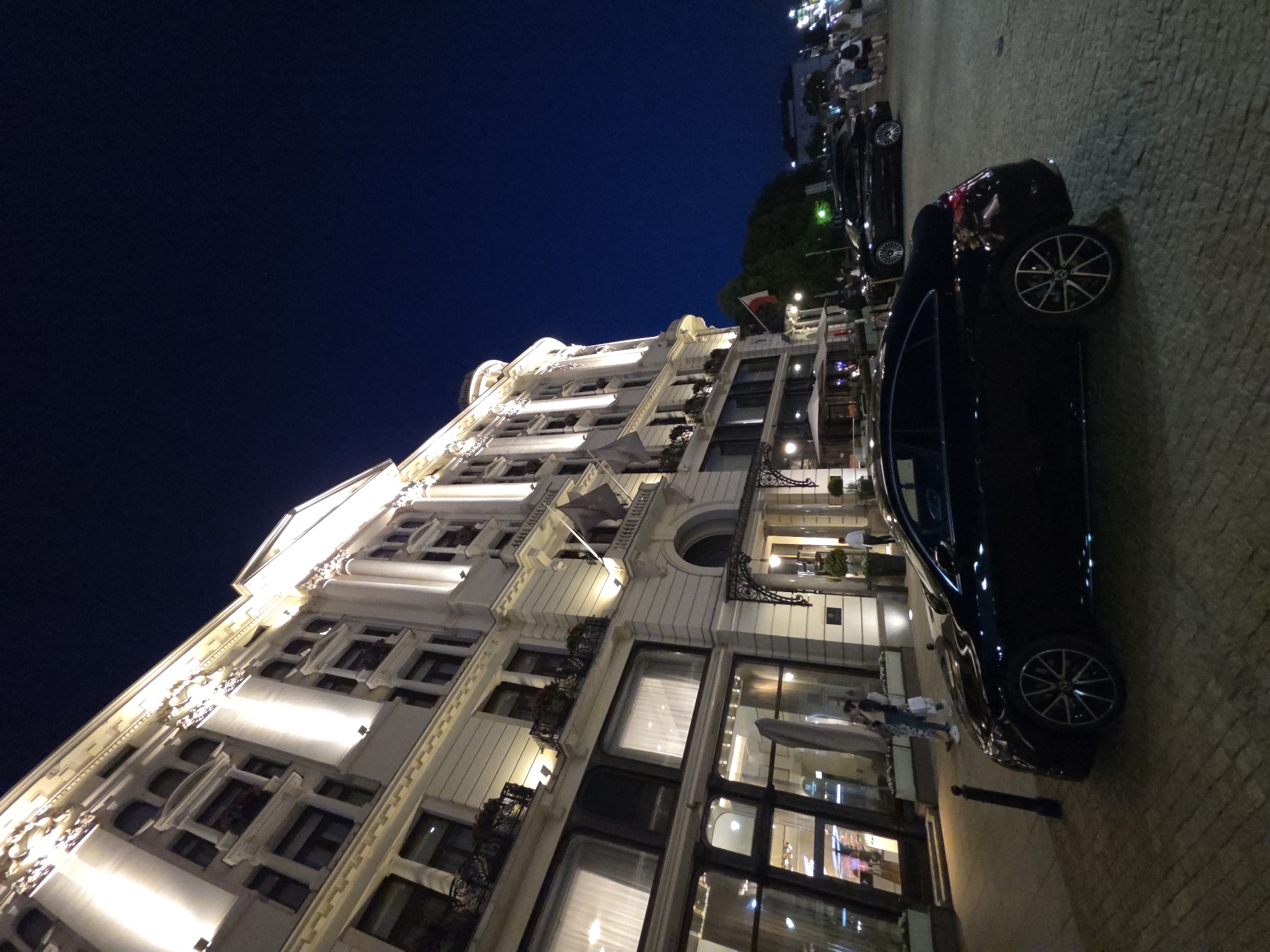 The image captures a nighttime scene in Warsaw, Poland, with a detailed view of a multi-story building and parked cars. The building is illuminated, showcasing its architectural features. A black car is parked along the side of the building. The sky is dark, indicating it is night. There are few people visible and the environment appears urban and well-lit.