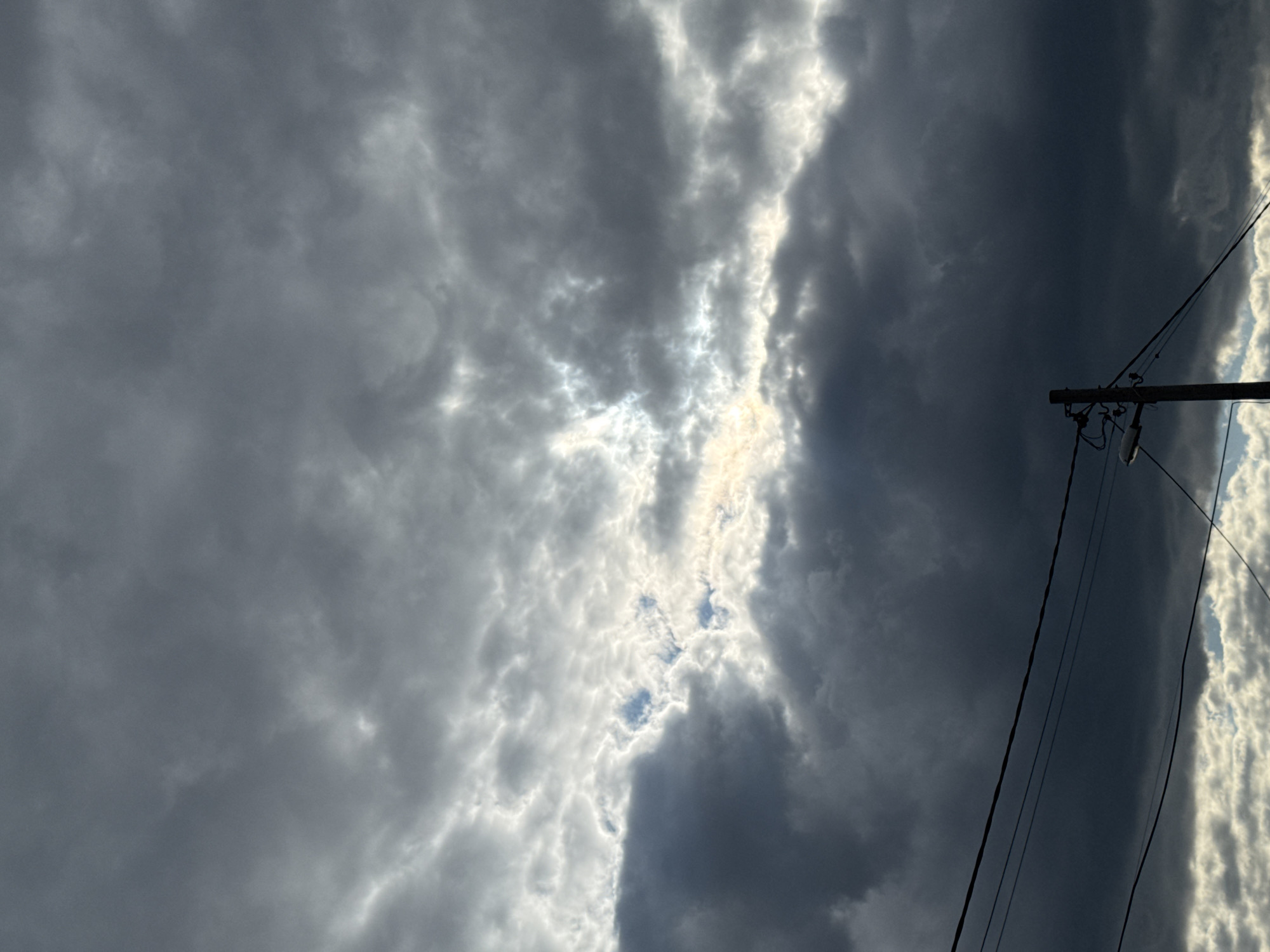 The image shows a view of the sky, likely in the city of Plovdiv, Bulgaria. Overcast conditions are evident, with dark and gray clouds dominating the scene. A break in the clouds allows some sunlight to penetrate, creating areas of brightness. A utility pole with attached wires is visible in the lower right portion of the image, suggesting an urban or suburban setting.