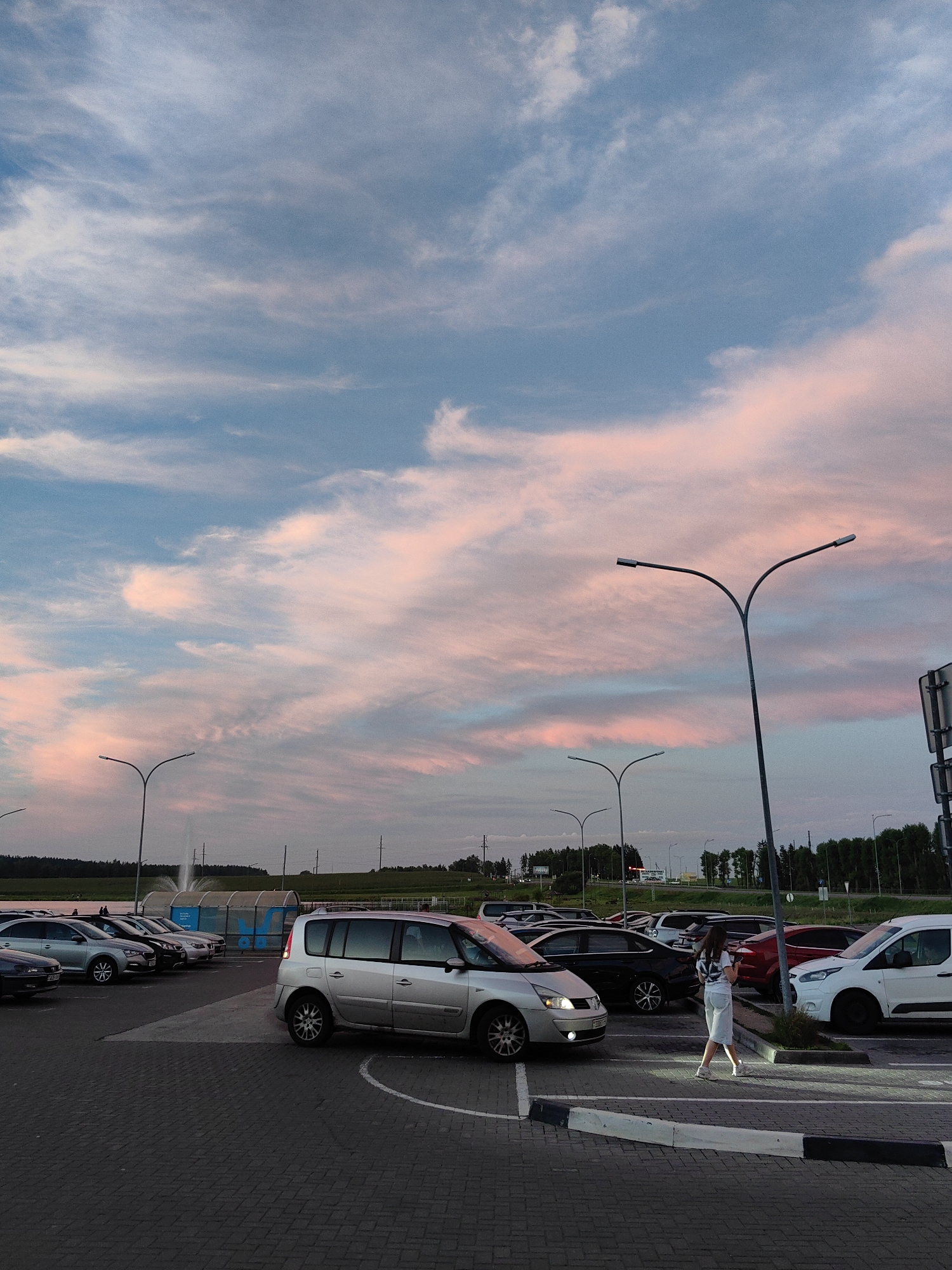 The image depicts a parking lot scene in Dzyarzhynsk, Belarus, during twilight. The sky is a prominent feature, filled with soft pink and blue clouds, indicative of sunset or sunrise. Several cars are parked in the lot, including a silver minivan in the foreground, a white van, and other vehicles. A woman wearing a white top and light-colored skirt is walking across the parking lot, holding a smartphone. Streetlights illuminate parts of the scene. In the background, there are trees and some infrastructure, including what appears to be a fountain or water feature behind a covered structure. The parking lot is paved with interlocking bricks.