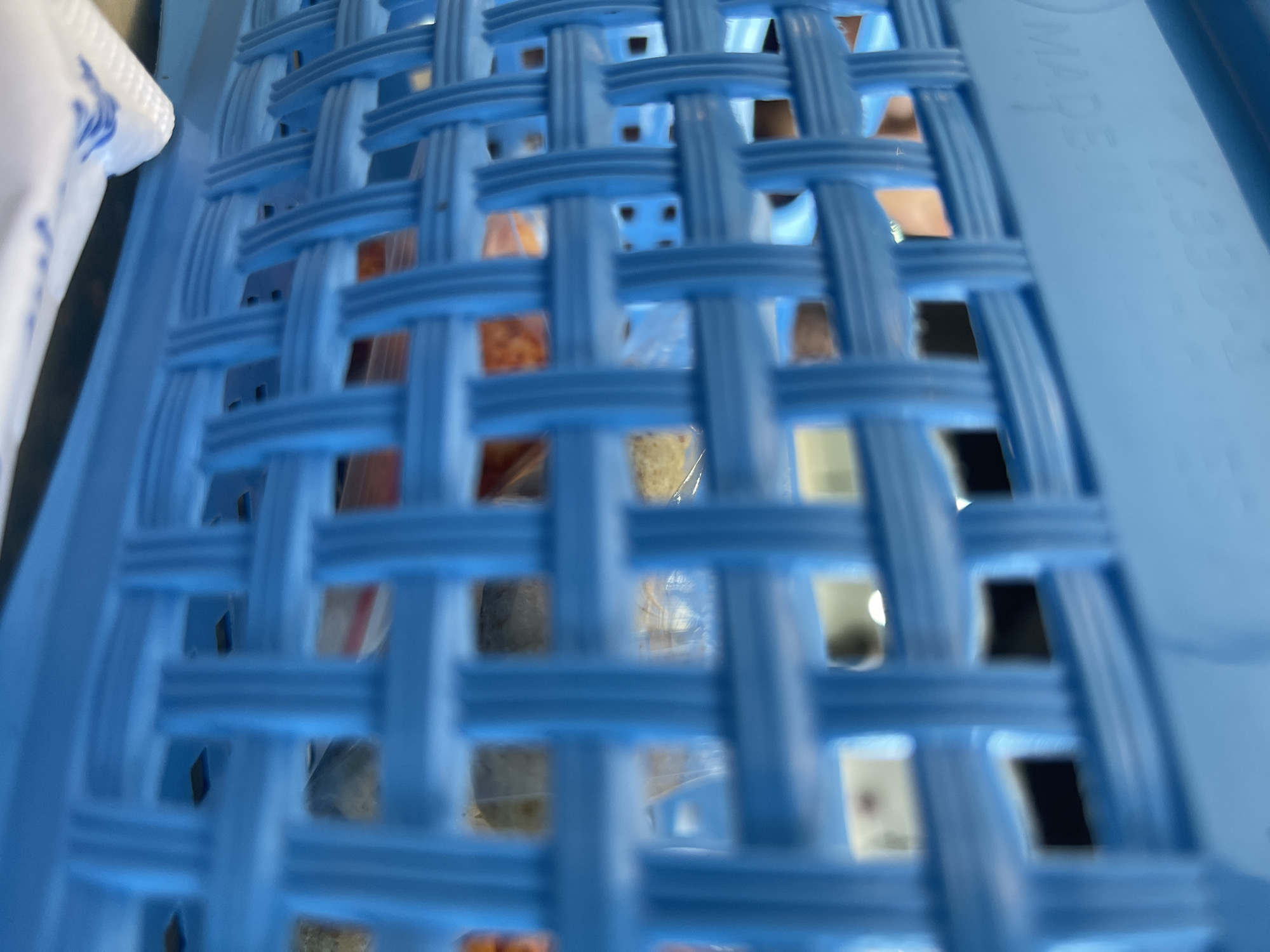 A close-up, slightly out-of-focus shot shows the woven texture of a blue plastic basket. Through the openings in the basket, indistinct items can be seen, some appearing to be food items in plastic packaging. The basket has embossed text, including "MADE" and what appears to be "K.933". The background is blurred, showing hints of indoor lighting and possibly other objects. The setting is not identifiable as Bireuenmeunasahdayah, Indonesia, based on visual cues in the image.