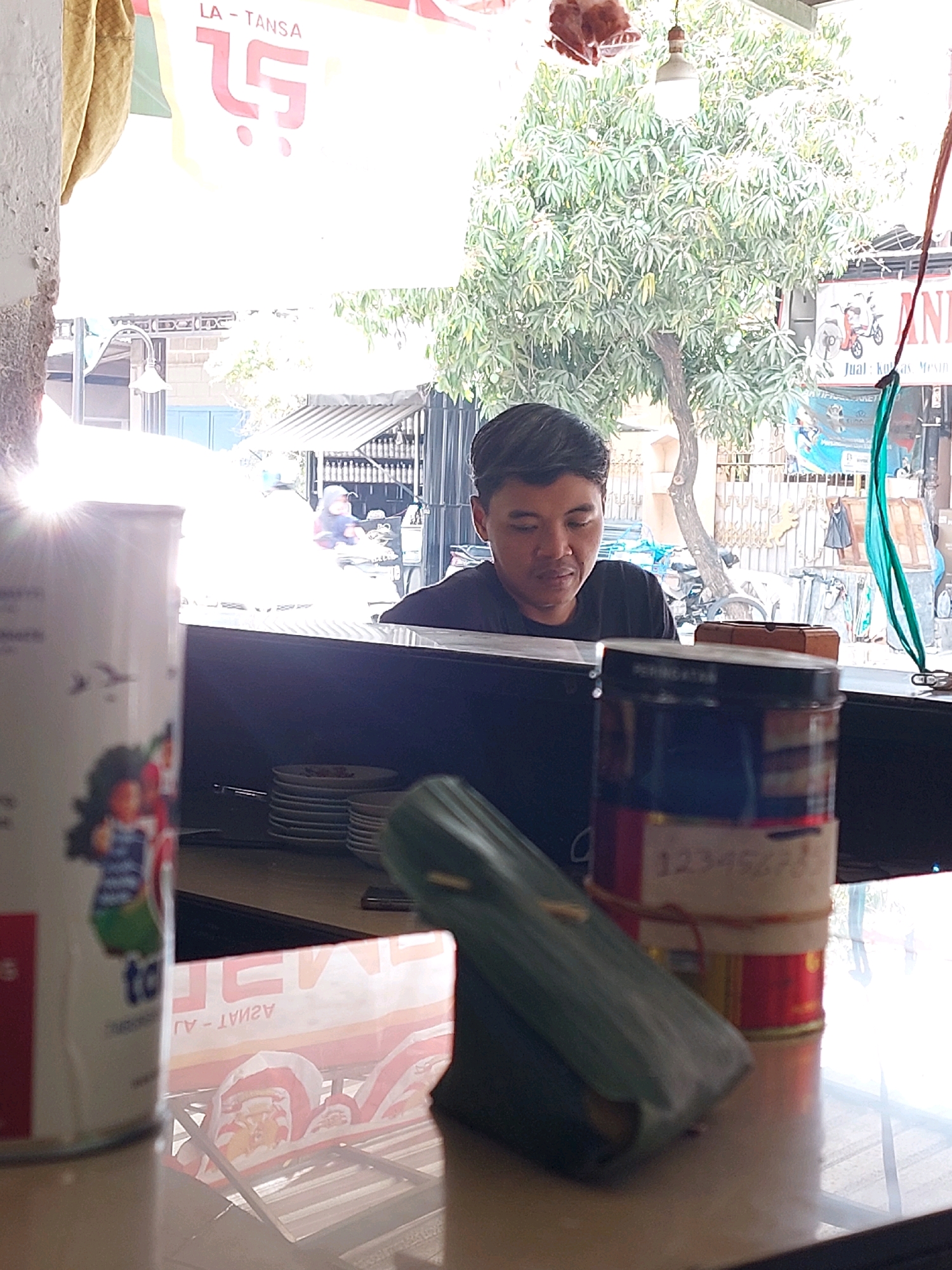 The image depicts a roadside food stall in Sumuragung, Indonesia. The stall appears to be set up in front of a building with a large banner displaying "LA-TANSA" and a stylized logo. A man is seated behind a counter, looking down at something in front of him. In the foreground, several items are visible on the counter, including a white cylindrical container with graphics and text, a stack of white plates, and a green, rectangular package. In the background, trees and other stalls are visible, suggesting a busy street scene. The lighting is bright, with sunlight streaming in from the left, creating a lens flare effect.