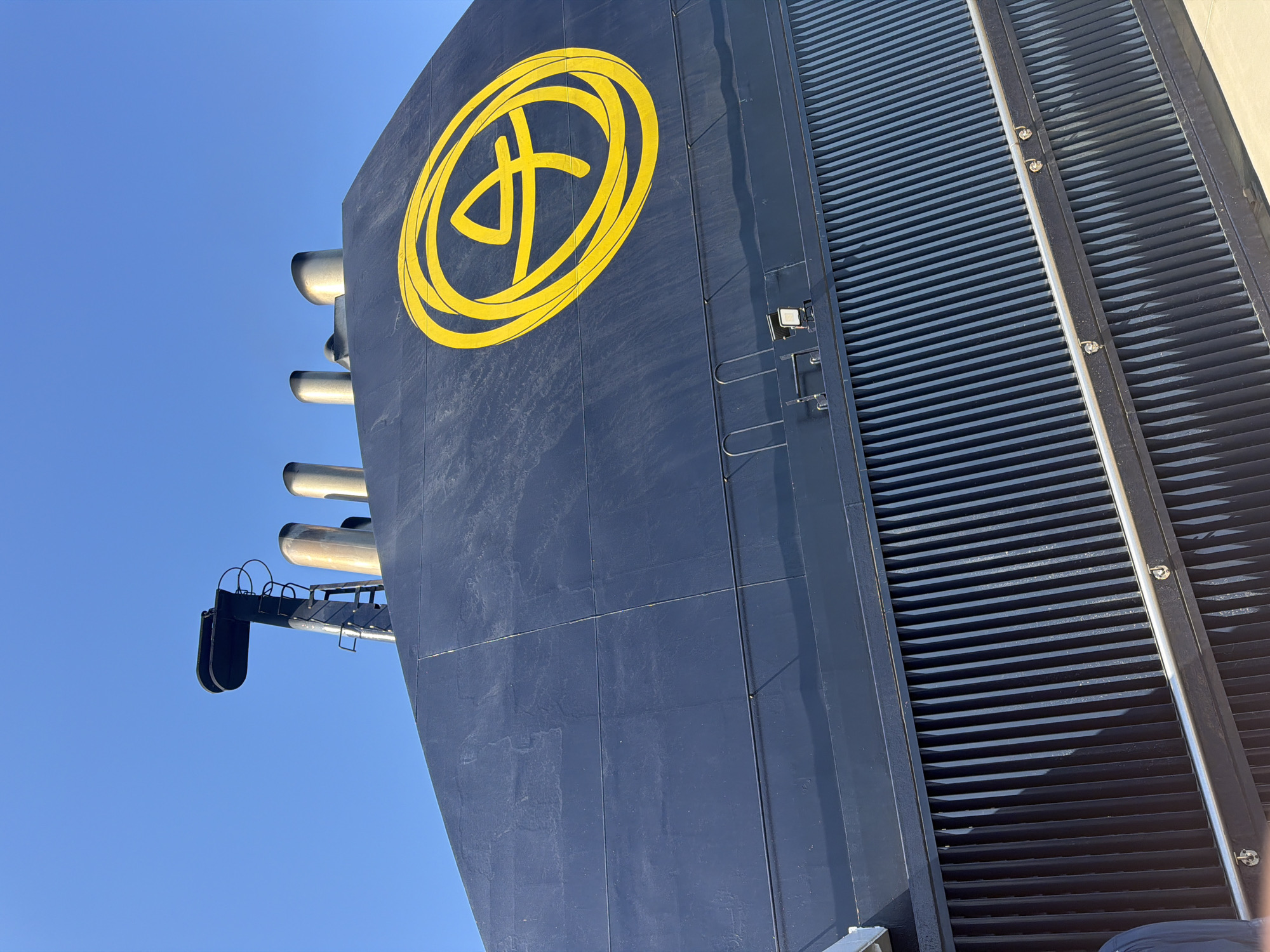 The image shows a close-up of the dark blue superstructure of a large vessel, likely a ferry, against a clear blue sky. Prominently displayed on the side of the vessel is a large, bright yellow circular logo containing abstract, stylized figures. Several metallic exhaust stacks emerge from the top of the superstructure, with a black aerial structure to the left. Below the main body of the superstructure is a section with vertical dark slats, suggesting ventilation or an outer hull component. A silver railing runs along the bottom portion of the image, crossing over the slatted section. The angle of the shot is from below, looking upwards, emphasizing the scale of the vessel. This vessel is docked at the port of Kastro - Kyllini, Greece, a location known for its ferry connections, particularly to the Ionian Islands. The presence of the ferry and its branding indicates it is a functional maritime transport in operation within this municipality.