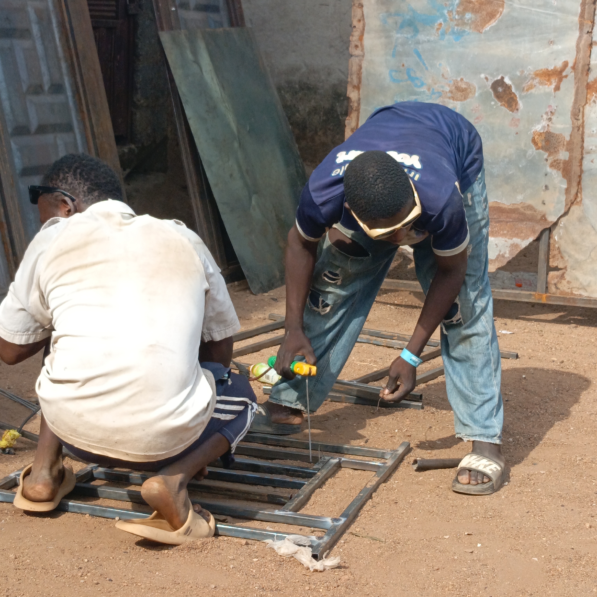 In Akwanga, Nigeria, two men are working on a metal frame on a dirt ground. One man wears a white shirt and is kneeling beside the frame. The other man, in a blue shirt and ripped jeans, leans over the frame, holding a yellow and green tool, while holding a piece of wire. Both men are wearing sandals. Metal sheets are stacked nearby. The background includes a weathered wall.
