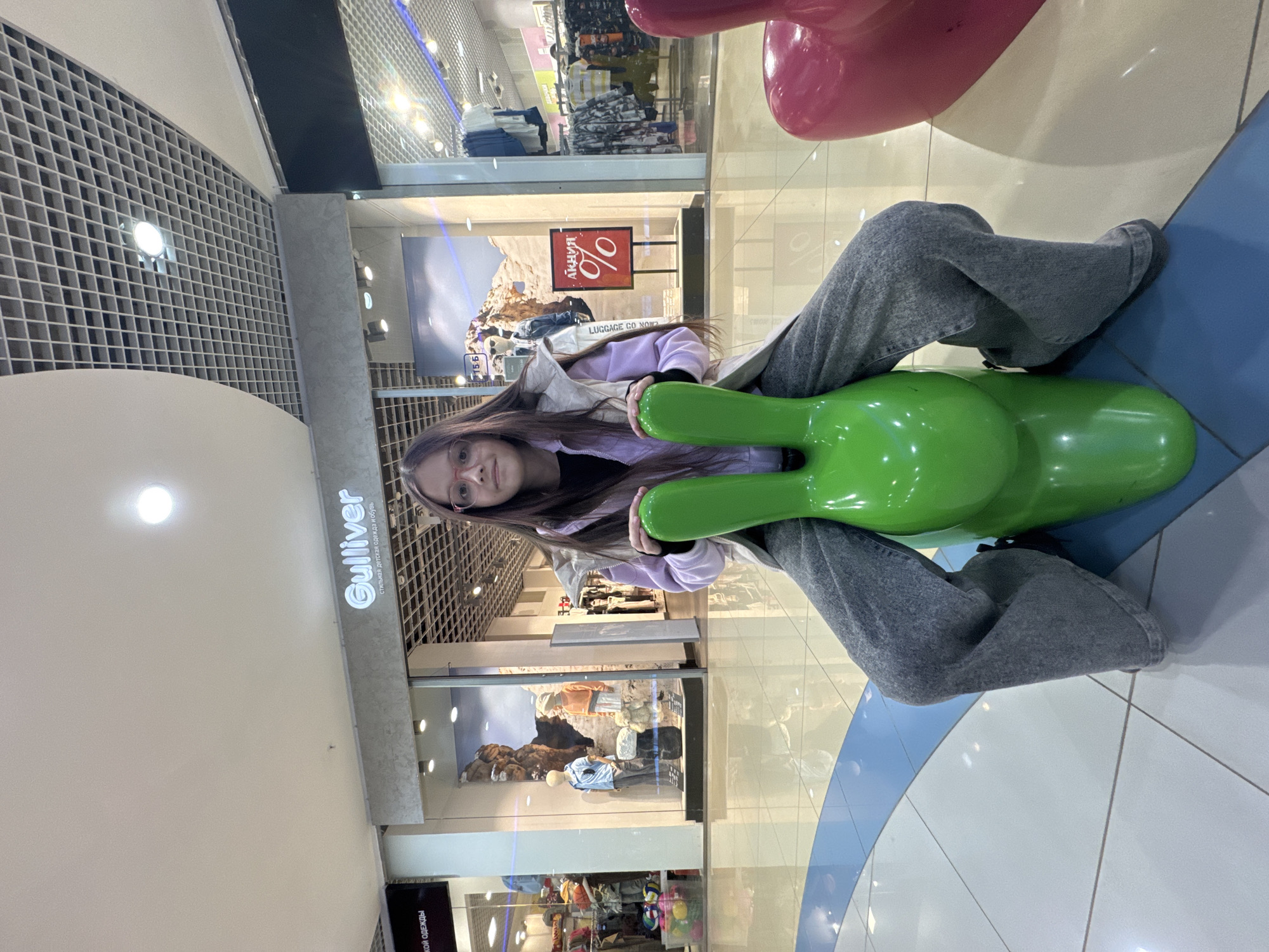 Here is a description of the image:
A young woman is seen at a shopping mall. She is sitting on a green, sculpted seat, positioned in front of a store with the sign "Gulliver" above the entrance. The store appears to sell children's clothing and shoes. The environment is well-lit, with a patterned ceiling and tiled floor. A red sign with "%" is visible in one of the store windows, indicating a sale. Other colorful sculpted seats are also present. The woman is wearing glasses, a lavender jacket, and gray pants.