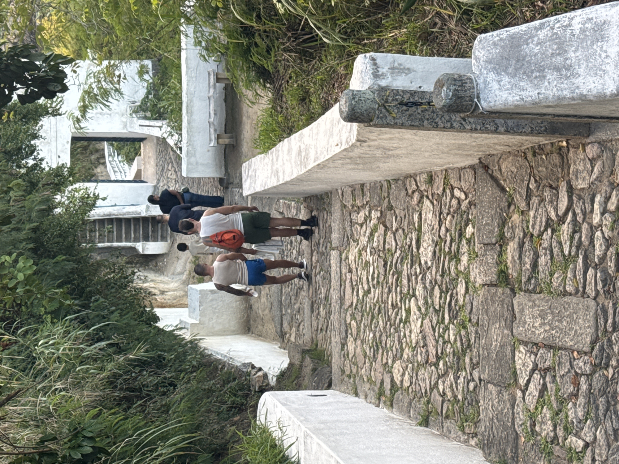 The image depicts a group of individuals walking up a stone-paved pathway lined with white stone walls and lush green foliage. Two men are in the foreground, facing away from the camera. The man on the left wears a white tank top and blue shorts, carrying a white towel over his shoulder. The man on the right wears a white tank top and green shorts, with an orange backpack. A third person, a child, is walking ahead of them, and two other individuals are further up the path, one wearing a black t-shirt and jeans, and another in a black top and jeans.
The pathway appears to be part of a historical or tourist site, possibly leading to an elevated area with stairs visible in the background. The architecture suggests a location in Brazil, potentially the Parque da Cidade in Niterói, known for its scenic views and historical structures. The environment is tropical, with dense vegetation and a warm, sunny atmosphere suggested by the lighting.