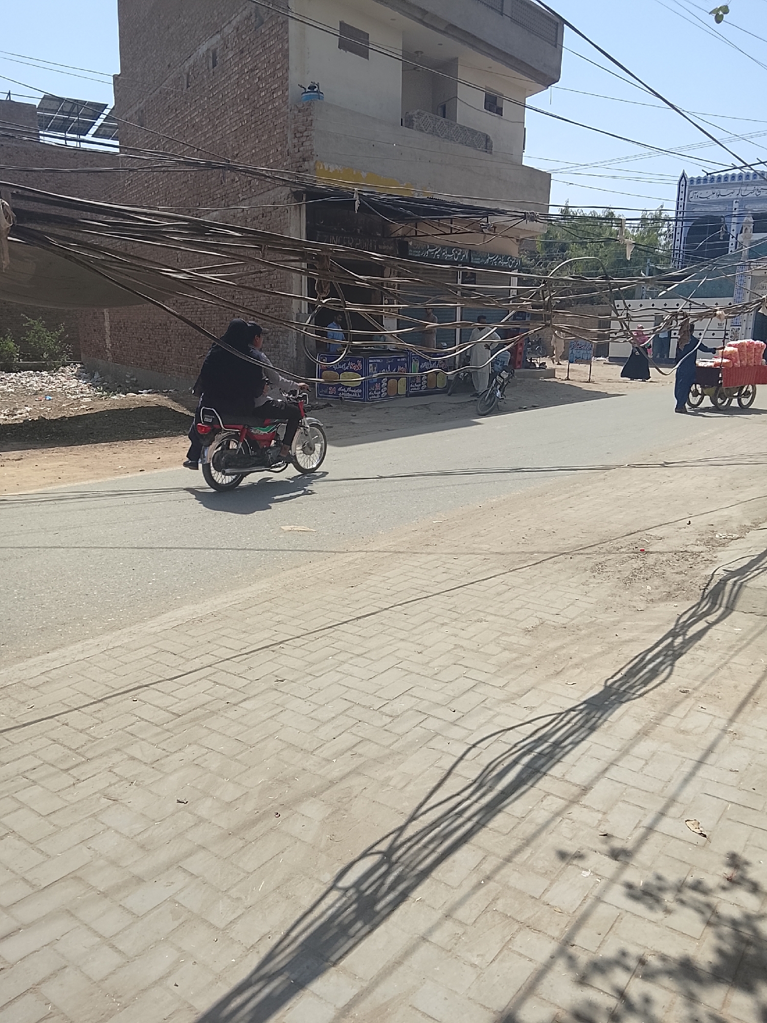 A medium-angle, full outdoor shot depicts a street scene during the daytime. The location is unknown. A man drives a motorcycle carrying a passenger down a paved street. There is an abundance of tangled power lines above the road, with the shadows of these lines visible on the brick-paved portion of the road. To the right of the motorcycle is a cart with red sacks. Buildings flank the road, and a person is visible walking in the distance.