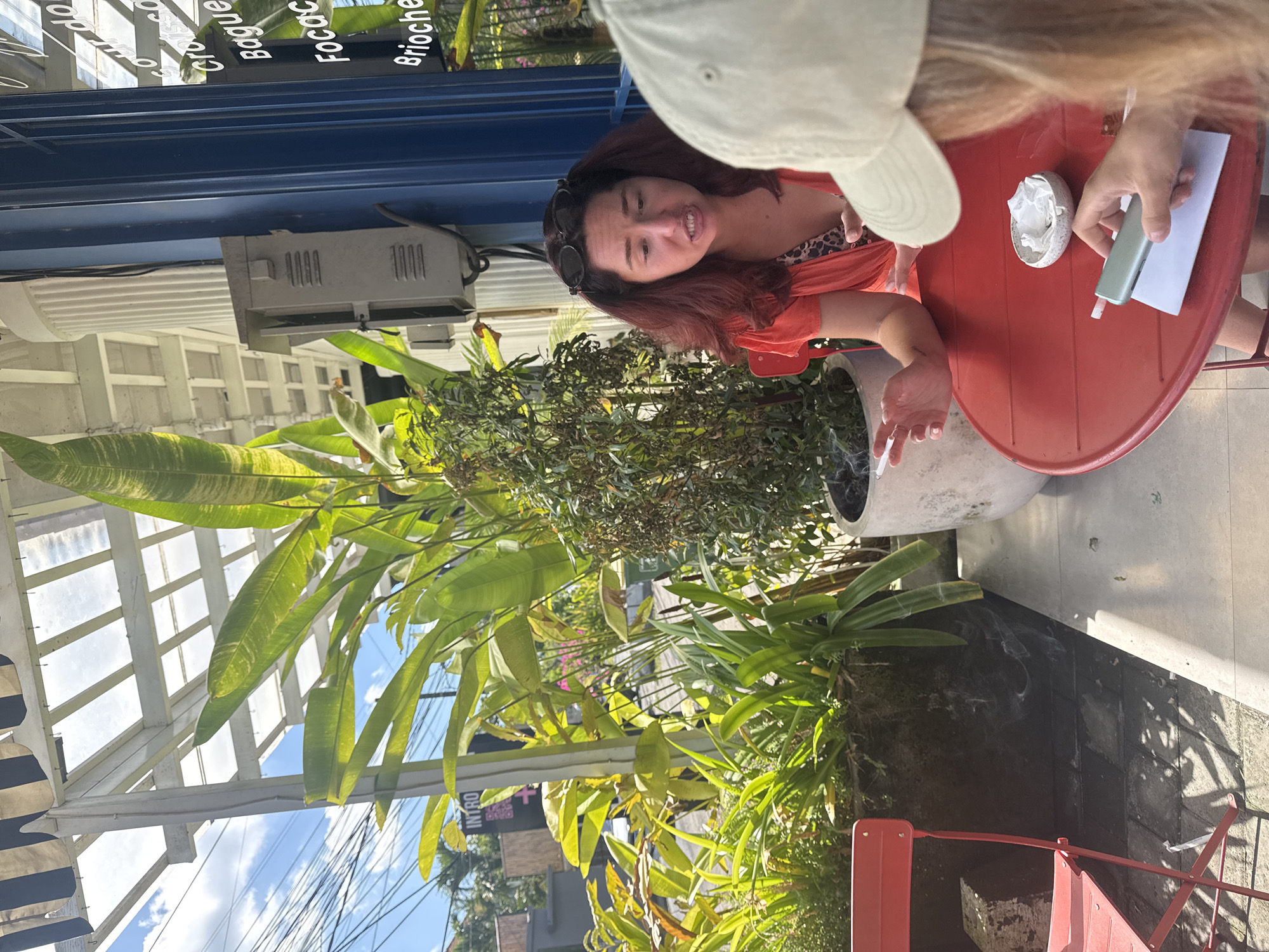 An outdoor scene captures two individuals at a red metal cafe table under bright daylight. The image is taken from a slightly elevated perspective, looking down, and appears rotated.
A woman with reddish-brown hair and sunglasses pushed up on her head is seated on the left, wearing an orange top over a leopard print garment. She is smiling and looking towards the camera, holding a lit cigarette in her left hand, with visible smoke swirling upwards. On the right, another person is partially visible, showing light-colored hair, a light-colored baseball cap, and an arm. This person holds a light green rectangular object, possibly a phone or vape, and a white paper over the table. A small white bowl filled with crumpled white paper or tissues sits between them on the table.
The setting is an outdoor cafe or restaurant, characterized by lush green tropical plants in pots and surrounding foliage, suggesting a warm climate. A large grey concrete planter is visible near the table. Overhead, a white slatted roof structure allows sunlight to filter through, revealing blue sky and clouds. A black and white striped awning is seen on the far left. Below a prominent blue horizontal beam, a grey electrical box is mounted. In the background, hints of a street or pathway with overhead wires are visible.
Visible text on a sign above the blue beam includes "Cro" (likely Croissant), "Baguet" (Baguette), "Focac" (Focaccia), and "Brioche," suggesting a bakery or cafe menu. The overall atmosphere appears casual and relaxed, indicative of a pleasant break in North Kuta, Indonesia, as suggested by the location context.