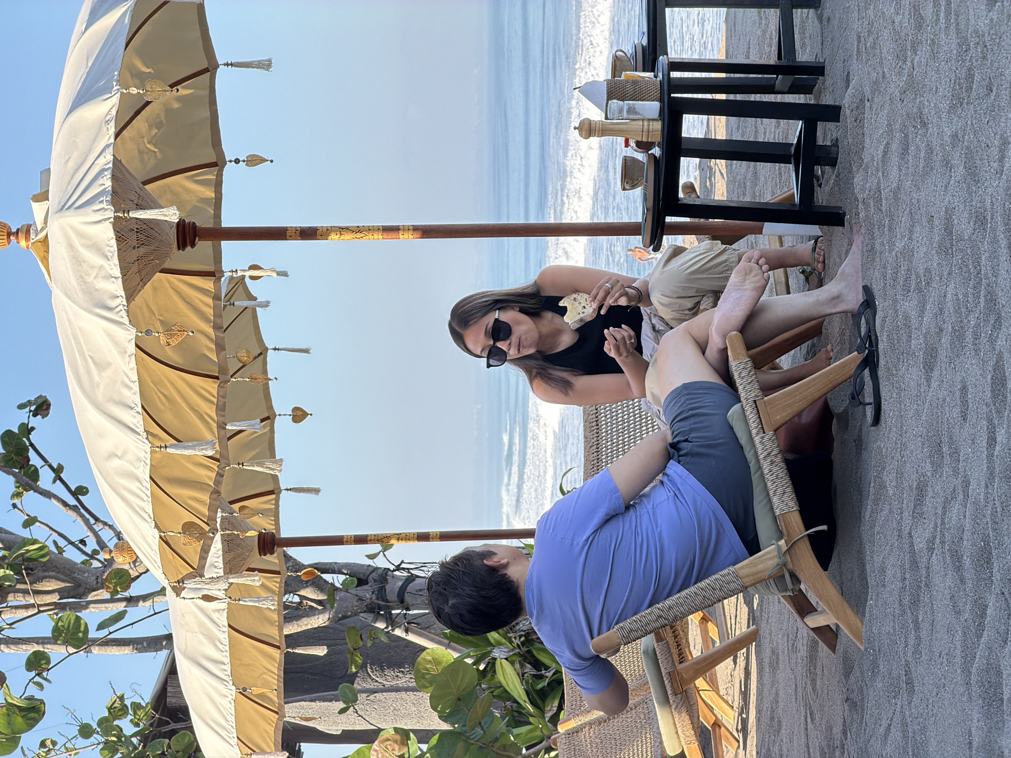Two individuals are seated on beach chairs on a sandy shore, facing the ocean under two large, decorative umbrellas. The man on the left, wearing a blue t-shirt and dark shorts, is looking out at the water. The woman on the right, wearing a black tank top and light-colored pants, is holding a piece of bread and appears to be eating. A small black table is positioned between them, holding two cups, salt and pepper shakers, and other items. The ocean with waves is visible in the background, under a clear blue sky. The scene is located in North Kuta, Indonesia.