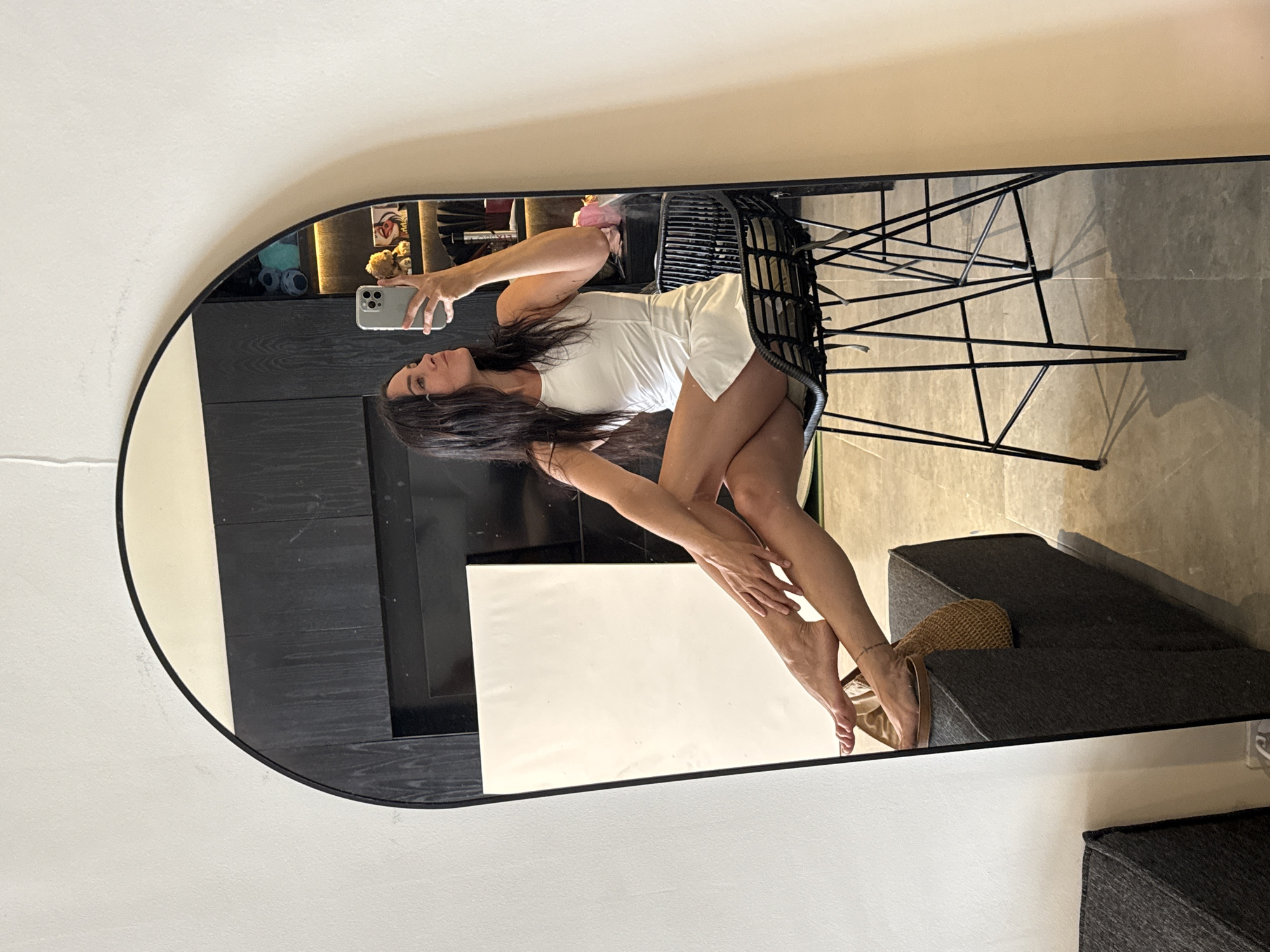 A woman with long, dark hair is taking a selfie in a large, arched mirror. She is wearing a white, sleeveless dress and gold heeled sandals. She is sitting on a black, metal-framed stool with a woven seat. Her legs are crossed, and her right foot is resting on a dark grey, rectangular ottoman covered in fabric. Her left foot is also on the ottoman. In her right hand, she holds a silver smartphone. The mirror reflects the woman, the stool, the ottoman, and a portion of the room behind her. The room appears to be modern, with dark wood cabinetry and shelves visible in the background. On the shelves, there are various decorative items, including stuffed animals and a bouquet of pink flowers. The floor is tiled. The image is taken indoors, likely in a residential setting in North Kuta, Indonesia.