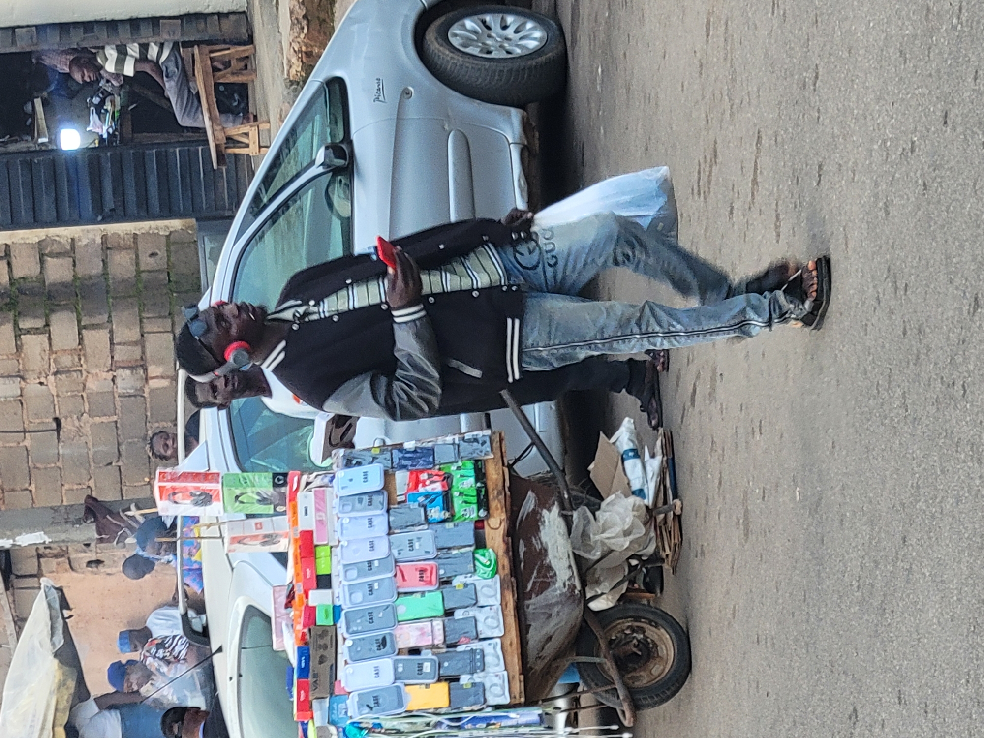 In Barakin Lerawi, Nigeria, a man walks past a mobile cart filled with various phone cases, while a silver car is parked nearby. The cart is laden with rows of phone cases, neatly displayed on its wooden surface. Behind the cart, a silver vehicle is visible, its presence suggesting it is either parked or moving slowly. The man, wearing headphones and holding a red mobile phone, is captured mid-stride. The scene appears to be a street or marketplace, with a brick wall and a shop in the background. The overall impression is of a bustling urban environment in Nigeria.