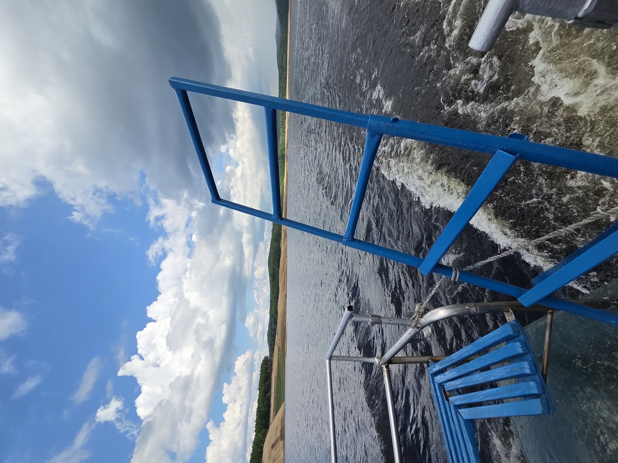 A bright, sunny day with scattered clouds is depicted, as seen from the stern of a boat moving through dark, choppy water. A blue metal ladder stands prominently in the foreground, angled towards the sky. To the left, a small, blue wooden bench is attached to the boat's railing. The water churns with a white wake behind the vessel. In the distance, a tree-lined shore with rolling hills stretches across the horizon, under a vast expanse of sky. The image appears to be taken at a reservoir or large lake, possibly in a rural or natural area.