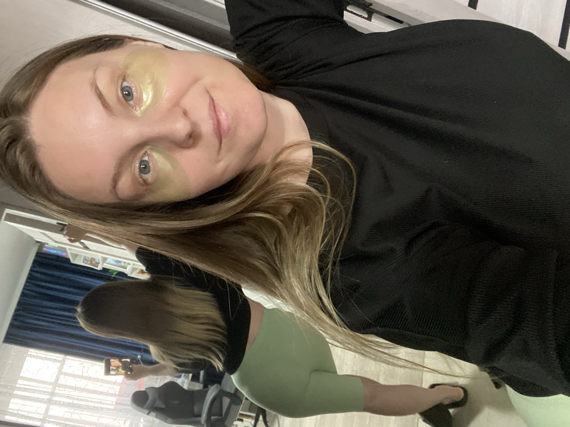 A woman is taking a selfie, wearing gold under-eye patches and a black shirt. In the background, a second woman is filming her with a smartphone. The second woman is wearing a black shirt and light green shorts. The background includes a window with blinds, a bookshelf, and blue curtains. The location is not discernible as Noviy, Russia.