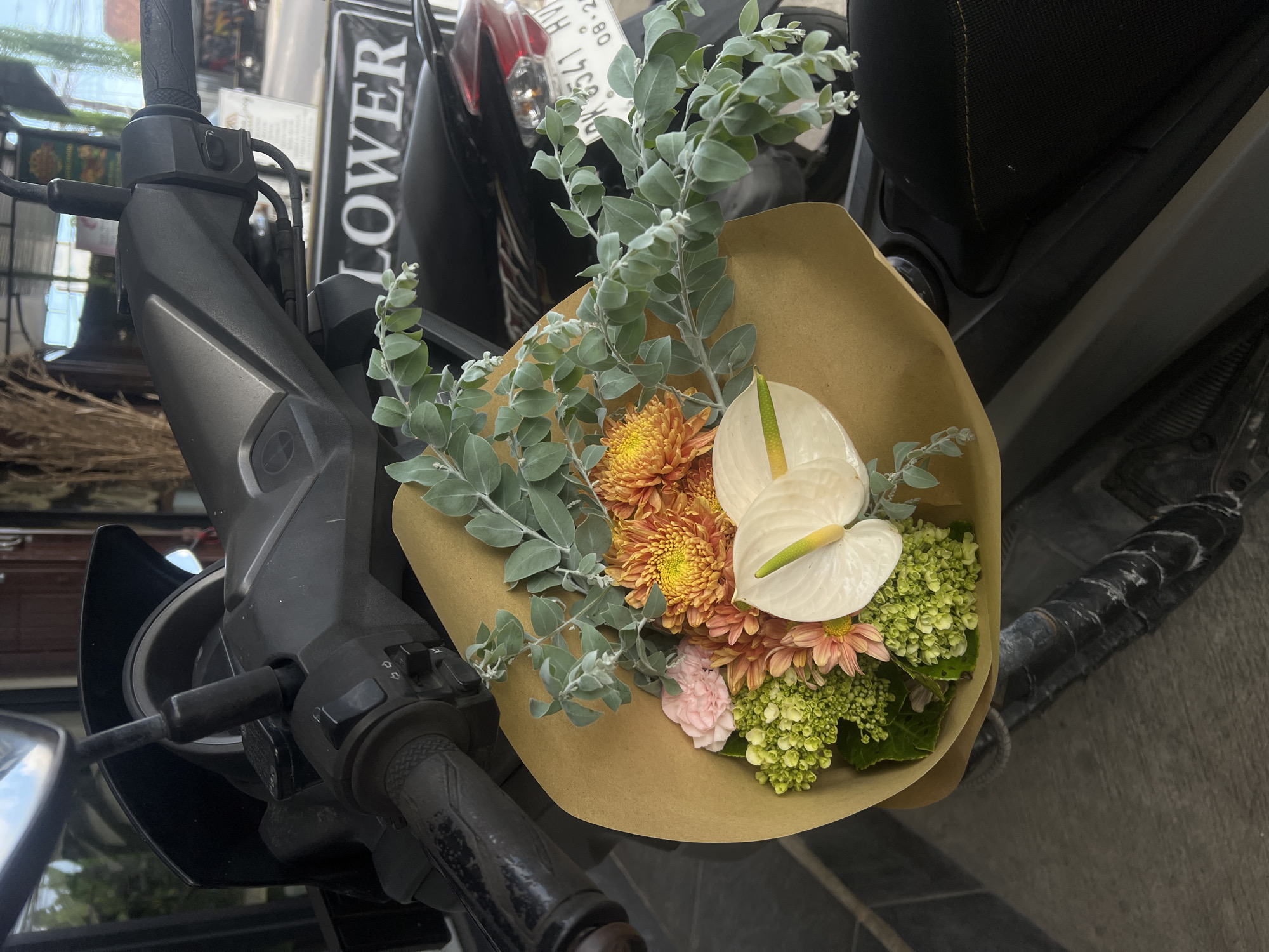 The image shows a bouquet of flowers placed on the handlebar of a gray scooter. The bouquet contains two white anthurium flowers, orange chrysanthemums, a pale pink carnation, and green hydrangeas, wrapped in brown paper. The scooter is parked on a street. A sign reading "LOWER" is partially visible in the background. The license plate "DK 6341 HV" is visible on the scooter. The scooter is in North Kuta, Indonesia.