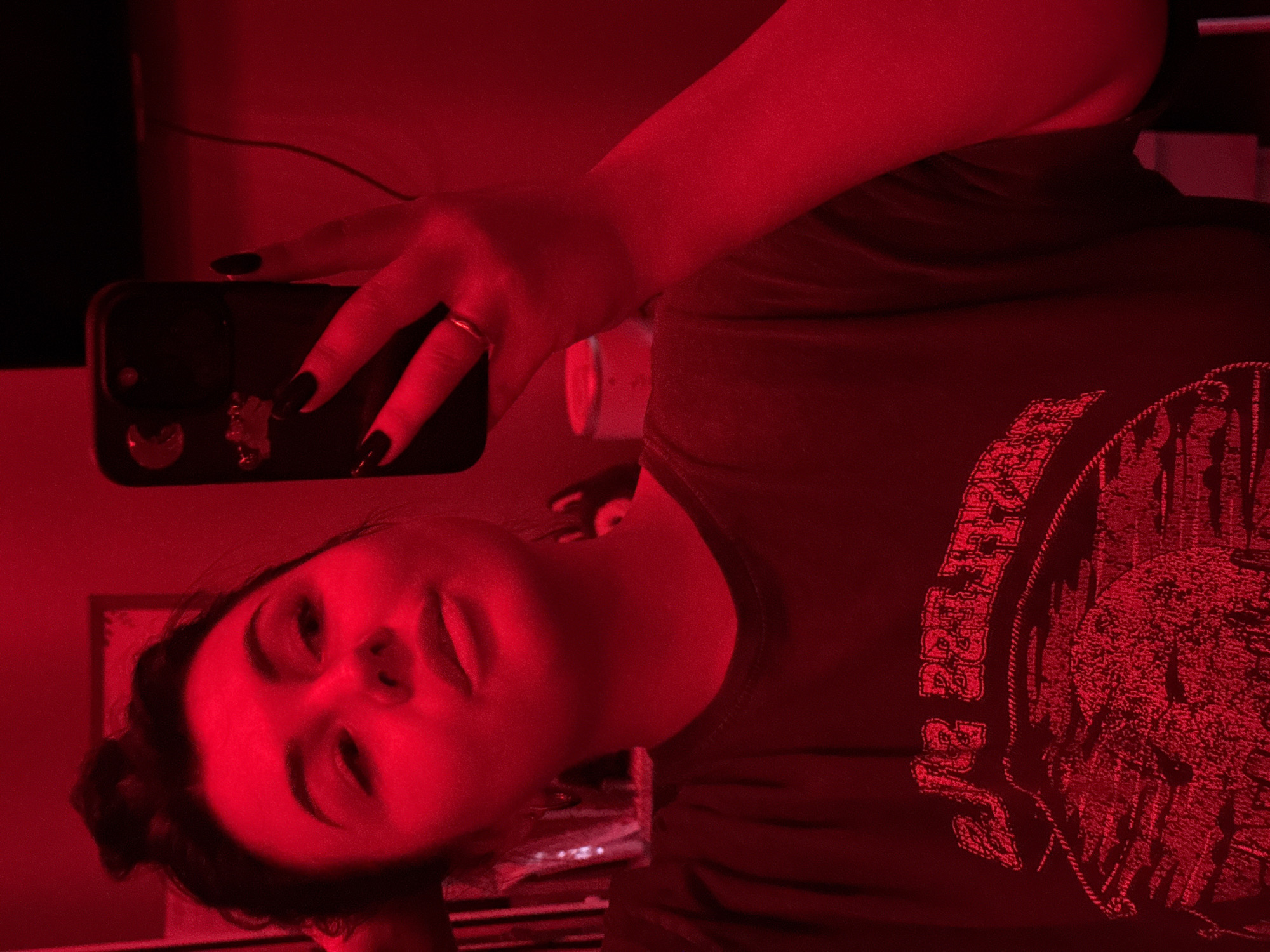 A woman with dark hair pulled back in a bun is taking a selfie in a room illuminated by red light. She is holding a black smartphone with a ring on her finger. She is wearing a dark T-shirt with a design on it. The background is blurred, but there appears to be a picture frame on the wall. There is no explicit information in the image to confirm or deny its location in Chaikovsky, Russia.