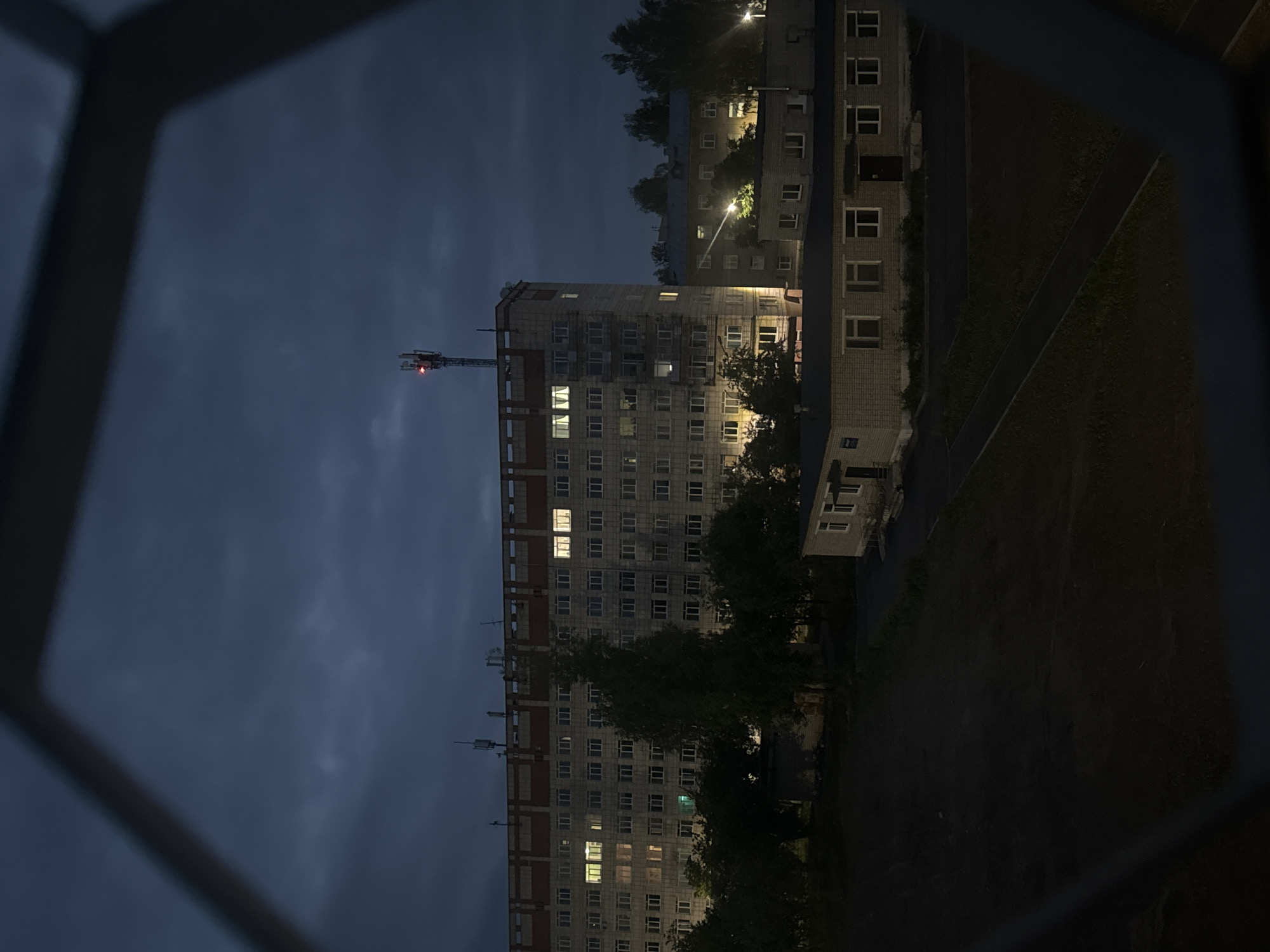 The image depicts an urban environment at dusk or night in Chaikovsky, Russia, viewed through a dark, angled frame that partially obscures the top and bottom edges. The scene is dominated by a large, multi-story, concrete-panelled building in the midground, featuring numerous evenly spaced windows. Multiple windows on different floors are illuminated with warm yellow light. A cellular tower with a red blinking obstruction light is prominently visible atop the building. To the right and in the foreground, several smaller, brick-clad buildings with illuminated windows and exterior lights are present. Dark foliage, including trees and bushes, is interspersed between and around the structures. The foreground consists of dark, unlit ground with a visible pathway. The sky is dark blue-grey and overcast. No individuals or discernible actions are present within the frame.