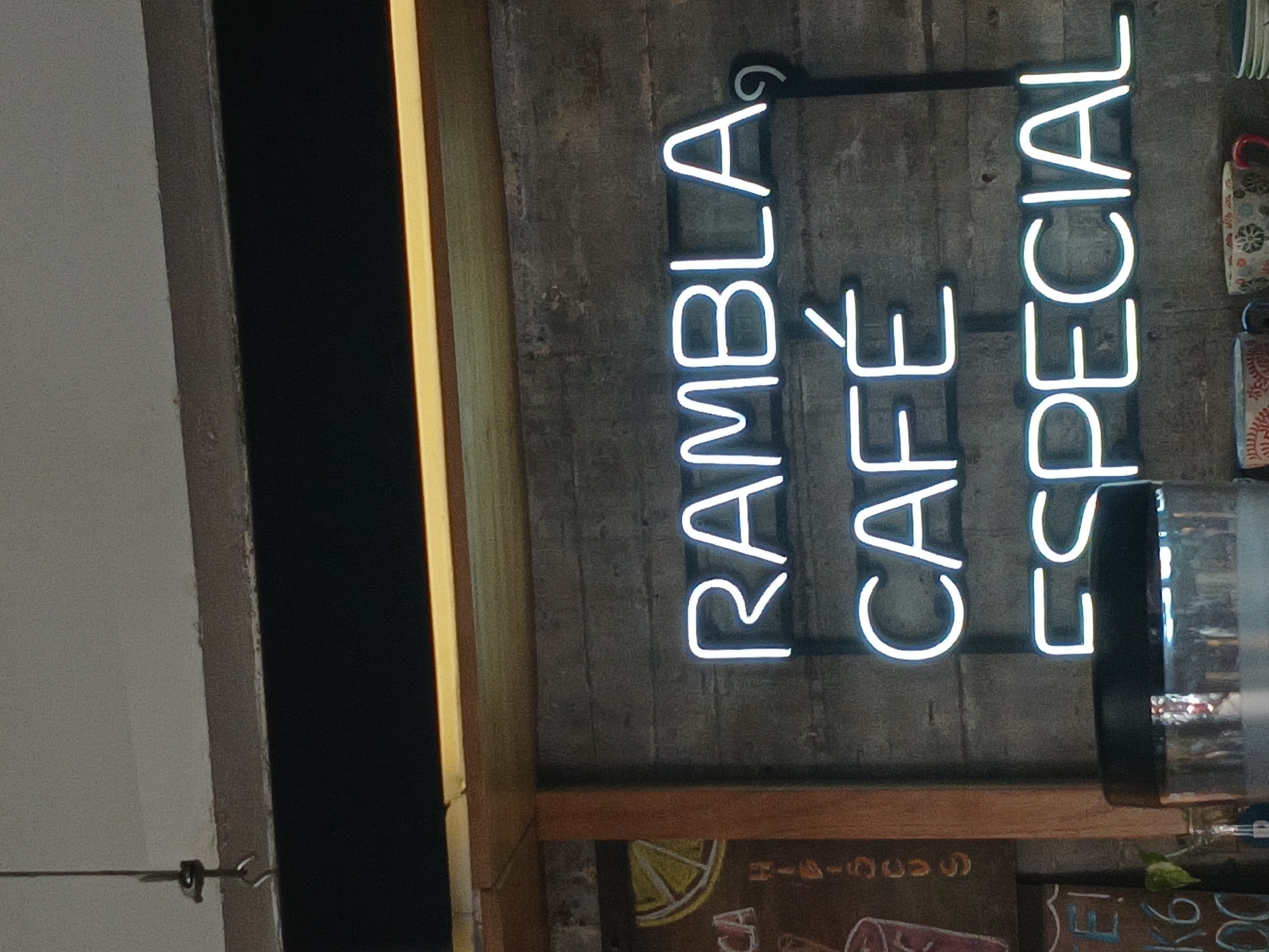 The image depicts a neon sign reading "RAMBLA CAFÉ ESPECIAL" vertically. The sign is illuminated in bright white and mounted on a textured gray wall. Below the sign, there are glimpses of other signage with illustrations including a lemon wedge and text partially visible. A glass container, possibly a dispenser or jar, partially obscures the right side of the sign. The overall setting suggests a cafe or restaurant. This image was taken in Buenos Aires, Argentina.