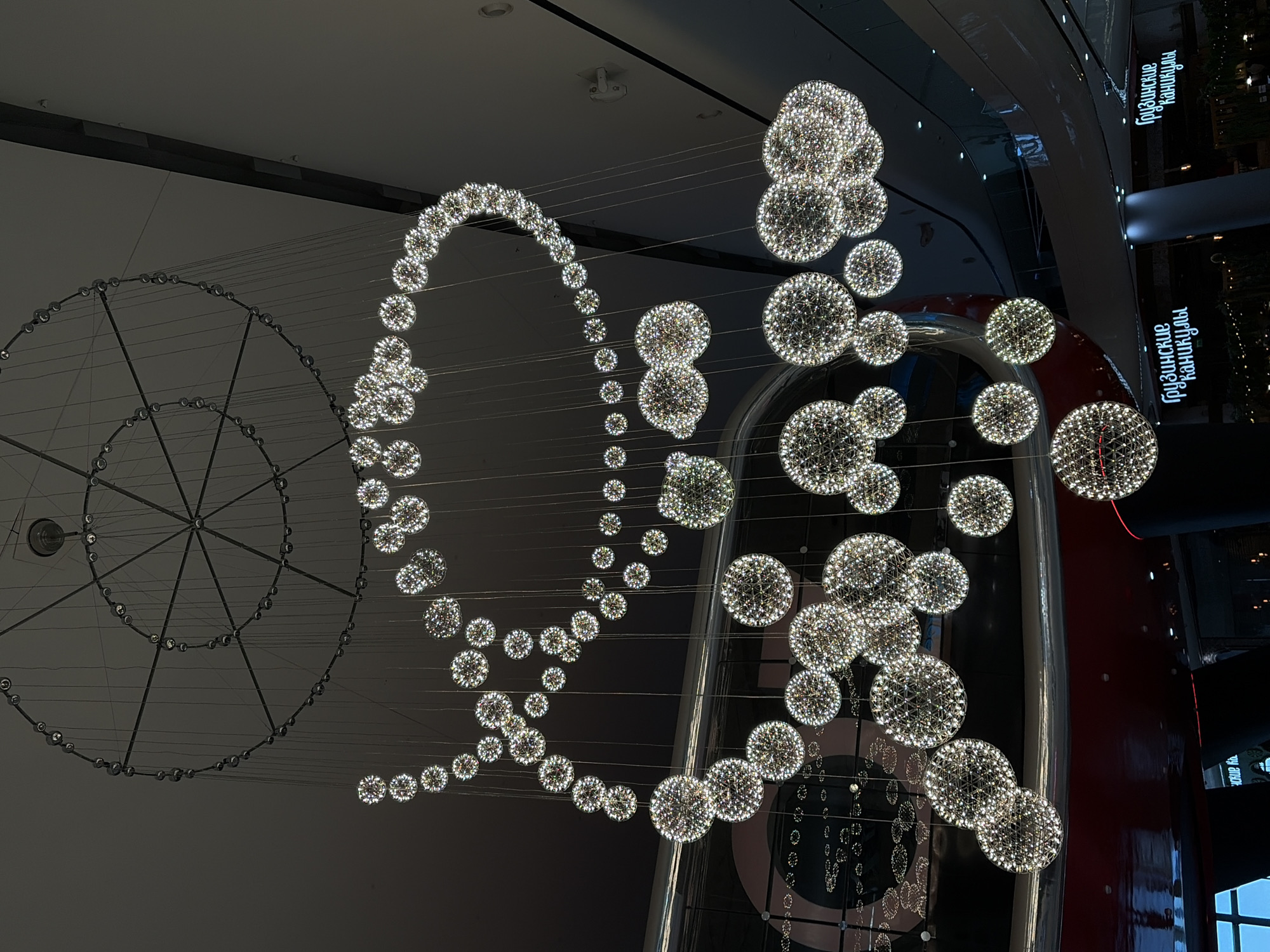 The image showcases a sophisticated indoor light installation within a modern building, likely a shopping mall in Krasnodar, Russia. The main subject is an elaborate arrangement of numerous sparkling spherical light fixtures suspended from a high ceiling. These spheres vary in size, composed of countless small, bright lights that create a shimmering, dazzling effect, and are arranged in an organic, flowing pattern that curves and cascades through the air.
To the left, a large, partially visible circular structure, resembling a skeletal Ferris wheel or a large ring, adds to the decorative elements. This structure is lined with smaller, unlit circular components and connected by thin wires. The background features dark, sleek architectural surfaces, including walls and possibly reflective ceilings, which contribute to the depth and modern aesthetic of the space. Some vertical architectural elements exhibit a reddish-brown color on the right.
The scene appears to be devoid of people, focusing solely on the grandeur of the architectural display. The controlled indoor lighting, with bright installations against a dark background, suggests either nighttime or an interior designed to highlight the illuminations.
Prominently displayed on a dark vertical surface on the right, visible multiple times and reflected, is Cyrillic text. The text reads "ПРИНЦИПИАЛЬНО НОВЫЙ," which translates to "FUNDAMENTALLY NEW" or "PRINCIPALLY NEW," likely a part of a store's branding or an advertising slogan within the commercial complex. The overall impression is one of modern elegance, artistic design, and perhaps festive decoration.