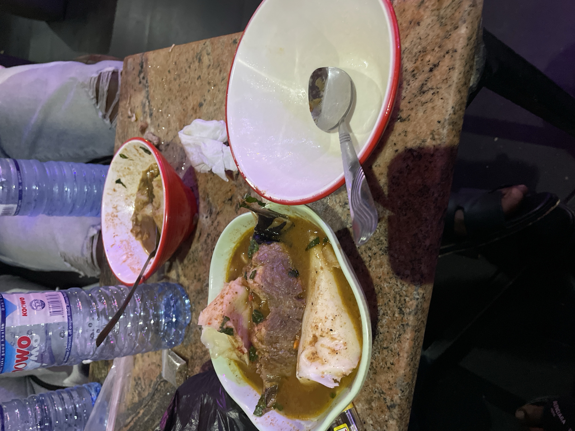 The image captures a scene in Obosi, Nigeria, where food remains are seen on a granite countertop. A near-empty bowl with a red rim and a spoon inside sits atop the counter, adjacent to a half-empty red bowl with a brown interior. Another bowl filled with soup containing large pieces of meat and what appears to be yam is placed nearby. Empty water bottles and a discarded tissue are scattered around the food. A person wearing sandals is visible in the periphery of the image. The environment appears to be dimly lit.
