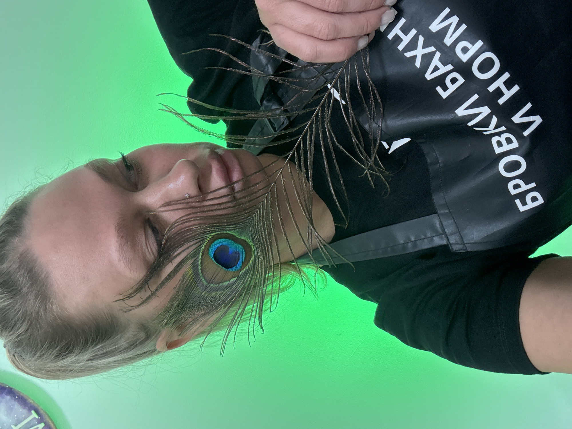 An artistically composed, close-up shot features a person, likely female, captured from the chest up against a vibrant, bright green background. The person's head is tilted, with eyes closed or looking downwards, conveying a serene expression. A small piercing is visible on their nose. Their hand holds a peacock feather, strategically placed across their face, with its striking iridescent blue-green "eye" positioned prominently near their cheekbone.
The individual is wearing a black garment, possibly an apron, which displays white Cyrillic text. When oriented correctly, the text reads "БРОВКИ БАХНУ И НОРМ", which translates to "I'll do eyebrows and it's fine" or "Eyebrows done and good." This text strongly suggests the person works in the beauty industry, likely as an eyebrow specialist, and the image may serve as promotional material. The uniform green background indicates an indoor setting, possibly a studio or salon. The lighting is uniform and bright, highlighting the subject and the feather.