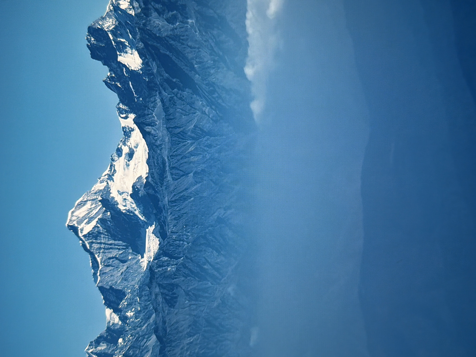 This is an image of a majestic mountain range under a clear blue sky. The mountains are snow-capped and rugged, with deep shadows and sharp ridges. In the foreground, a layer of clouds or mist obscures the lower slopes, creating a sense of depth and atmosphere. There are no people, objects, or actions depicted in the scene. The image is a landscape photograph, likely taken from a high vantage point. This image is not related to Kyiv, Ukraine.