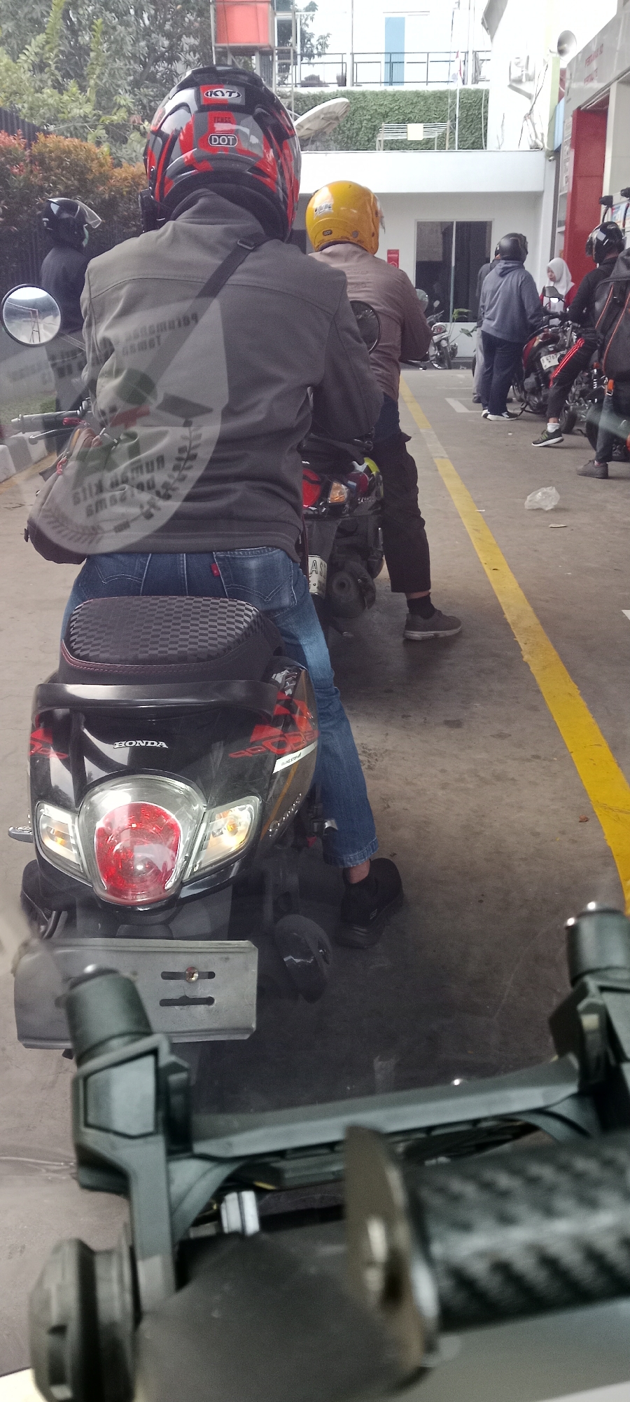 A motorcycle driver wearing a Kyt helmet and a gray jacket is seen from behind, riding a black Honda scooter. The driver is wearing blue jeans and black shoes. Another person wearing a yellow helmet is visible on a separate motorcycle behind the first driver. In the background, other motorcycles and people are gathered, possibly at a gas station or a busy street. The location is identified as Tangerang, Indonesia. The scene is captured from a vehicle in front of the first motorcycle.
