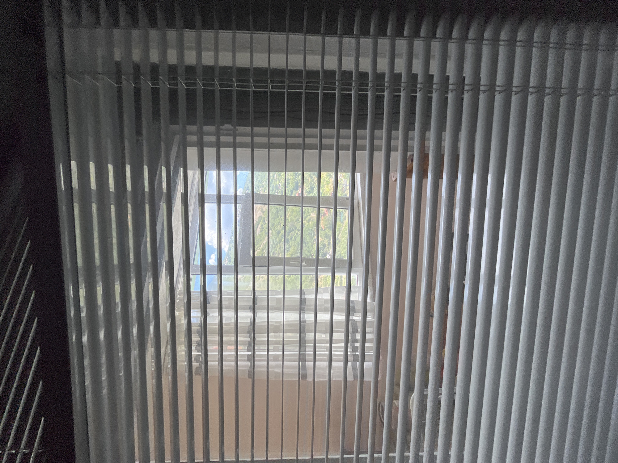 The image shows a view through horizontal blinds, looking out a window. The blinds are light gray and partially obscure the scene outside. Through the gaps in the blinds, a window with white sheer curtains can be seen. Beyond the curtains, a green, tree-covered hillside with some buildings is visible under a partly cloudy blue sky. The interior of the room is dimly lit, with a hint of a light-colored wall and some indistinct objects in the foreground, possibly furniture. The location in the city of Sopot, Bulgaria is not discernible from the image itself.