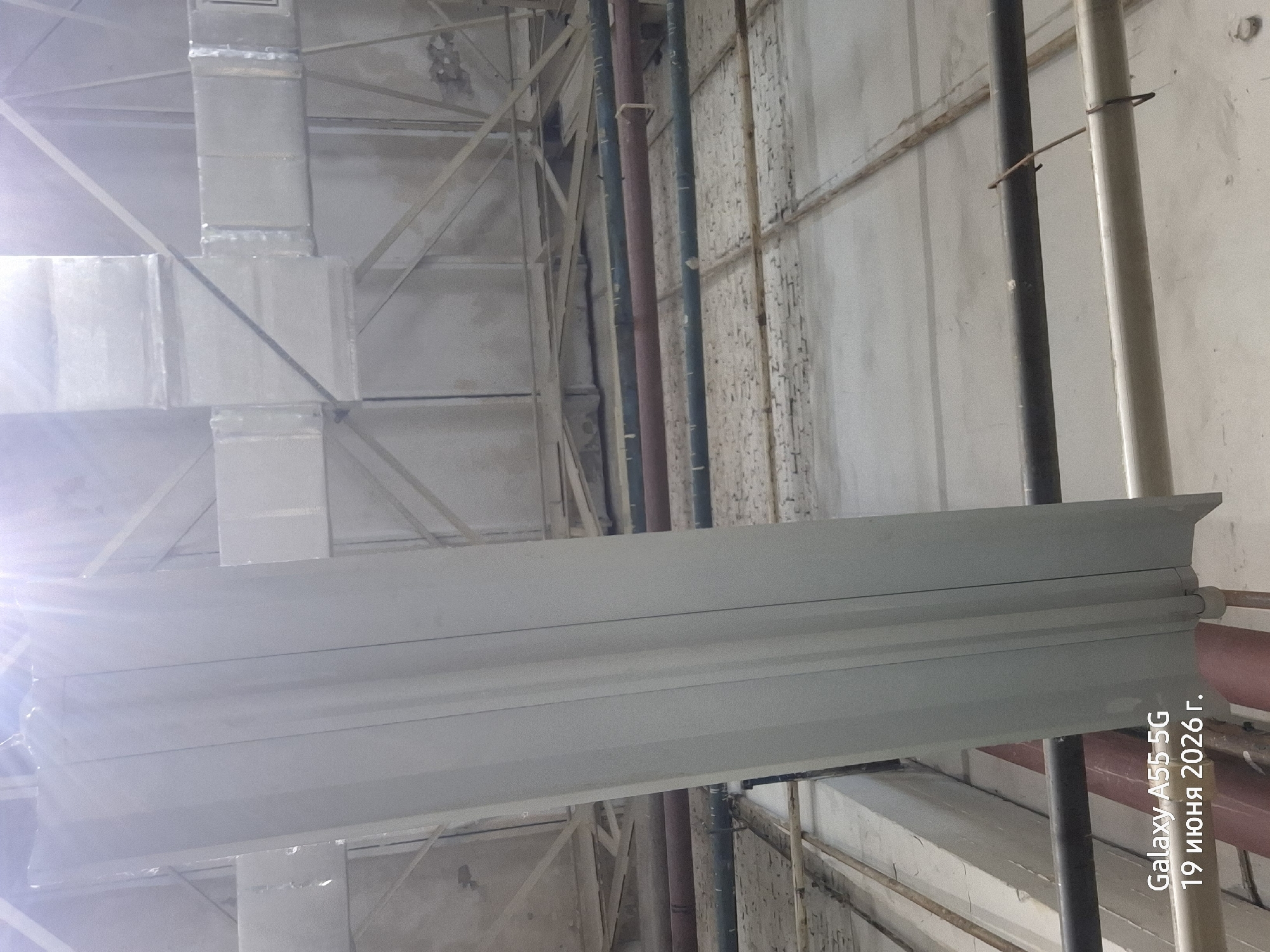 The image is a low-angle shot of the interior of a large, industrial-looking space, possibly a warehouse or factory. The ceiling is a complex network of pipes, ventilation ducts, and structural beams. A large, light-colored, vertical panel is prominently featured in the foreground, running from the bottom of the frame towards the top. This panel has a fluted or ribbed surface. Several horizontal pipes, some dark gray and some reddish-brown, are visible running across the mid-ground and background. The walls and ceiling appear to be constructed of concrete and metal. The overall lighting is somewhat diffuse, with a bright light source at the top of the image, suggesting overhead lighting or sunlight. In the bottom left corner, there is a watermark that reads "Galaxy A55 5G 19 июня 2026 г.", indicating the device and date the photo was taken, with the date in Russian. There are no people or animals visible in the image. The environment appears to be dusty and somewhat neglected. Based on the visible information, it is not possible to determine the specific location in Rostov-on-Don, Russia.