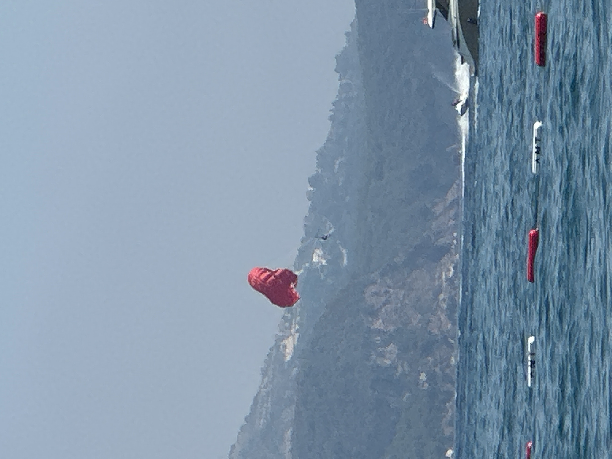 In the waters off Boreti, Montenegro, a parasailer is suspended in the air, their red parachute a vibrant contrast against the hazy blue sky and the lush, tree-covered slopes of the distant hills. Below, the azure sea is dotted with buoys, some red and some white. A sleek white yacht cruises on the right side of the frame, leaving a white wake in its path, while a personal watercraft speeds across the water in the foreground, its spray creating a misty trail. The overall scene depicts a lively summer day on the Adriatic coast, with various water activities taking place.
