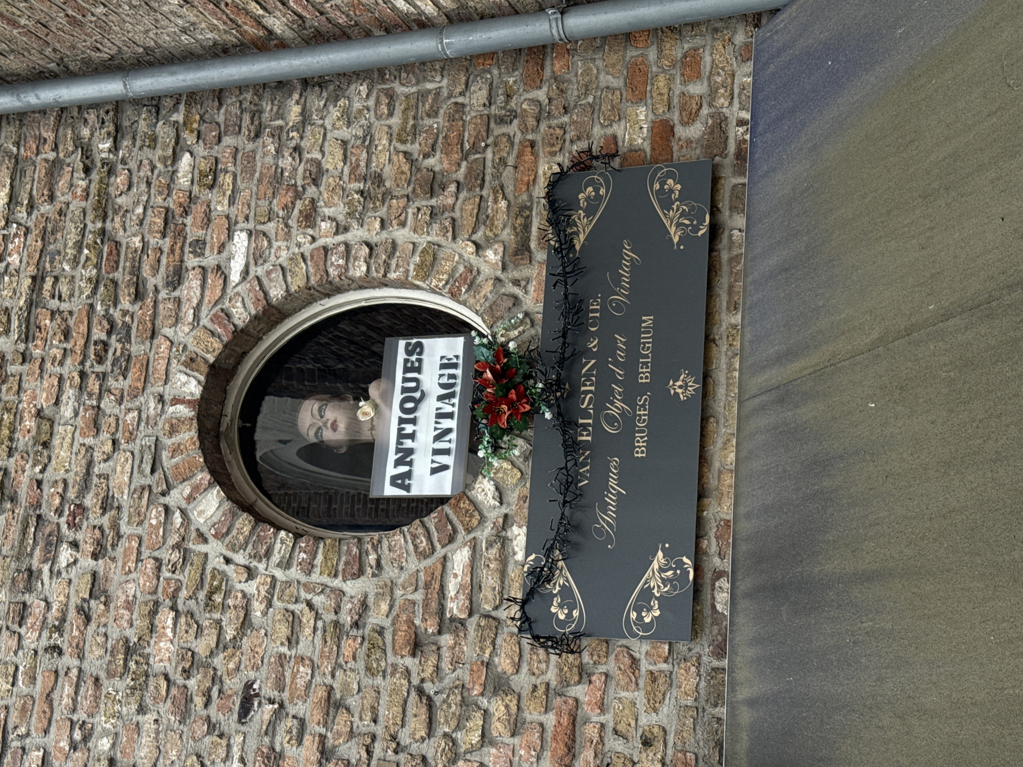 A mannequin head with red lipstick and a rose on its cheek is visible through a round window in a brick wall. A sign below the window reads "VAN ELSEN & CIE. Antiques Objet d'art Vintage BRUGES, BELGIUM". The sign is decorated with a black garland and gold scrollwork. A downspout runs along the right side of the brick wall. The bottom of the image shows a dark awning.