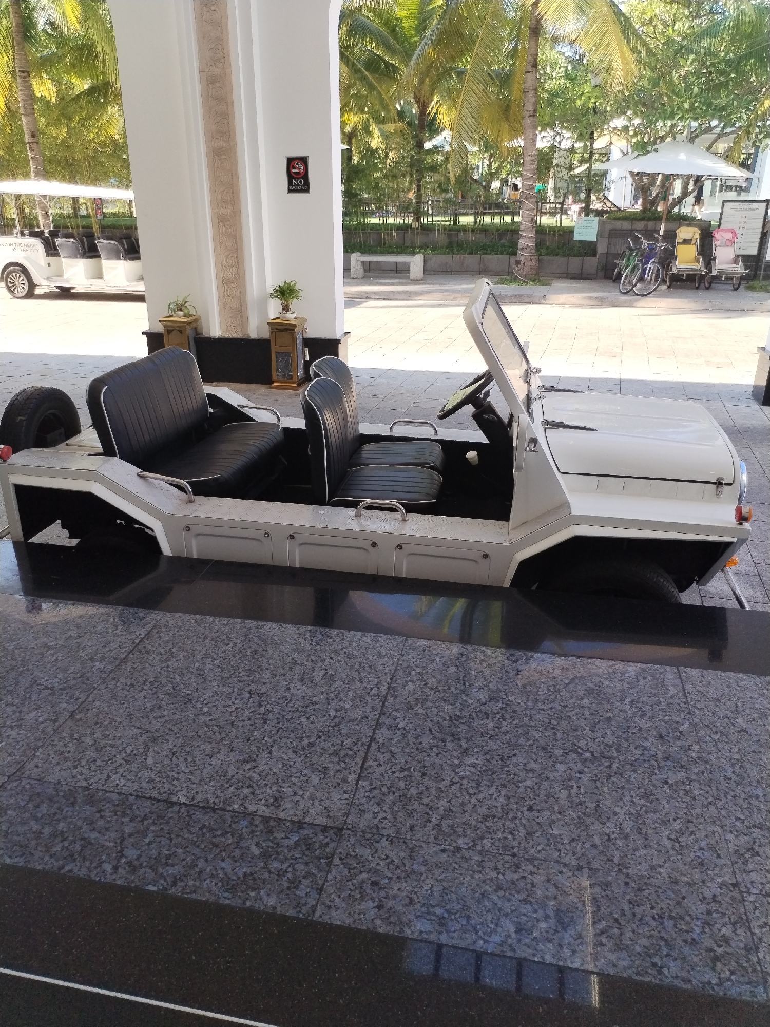 A white, open-top, four-wheeled vehicle, resembling a jeep or golf cart, is parked on a dark, reflective tiled floor. The vehicle has black vinyl seats with white piping, a steering wheel, and an open windshield. Behind the vehicle, palm trees and other greenery are visible, along with a white trolley or shuttle bus. A sign with "NO SMOKING" is displayed on a white pillar. The scene appears to be in an outdoor area, possibly a resort or hotel entrance, given the presence of the shuttle and manicured landscaping.