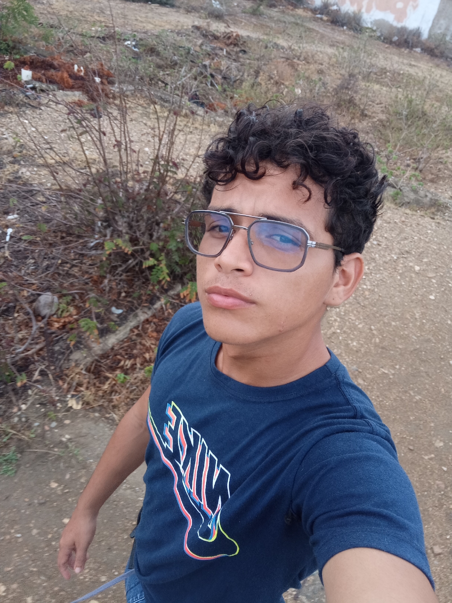 The image shows a young person, with curly dark hair, wearing glasses and a dark blue t-shirt with a colorful logo on the chest. They are standing outdoors in an arid, unpaved area with sparse vegetation and scattered debris. The background appears to be a hillside or embankment with some lighter colored structures or graffiti visible at the top. The lighting suggests it is daytime. The location is specified as Juan Griego, Venezuela.