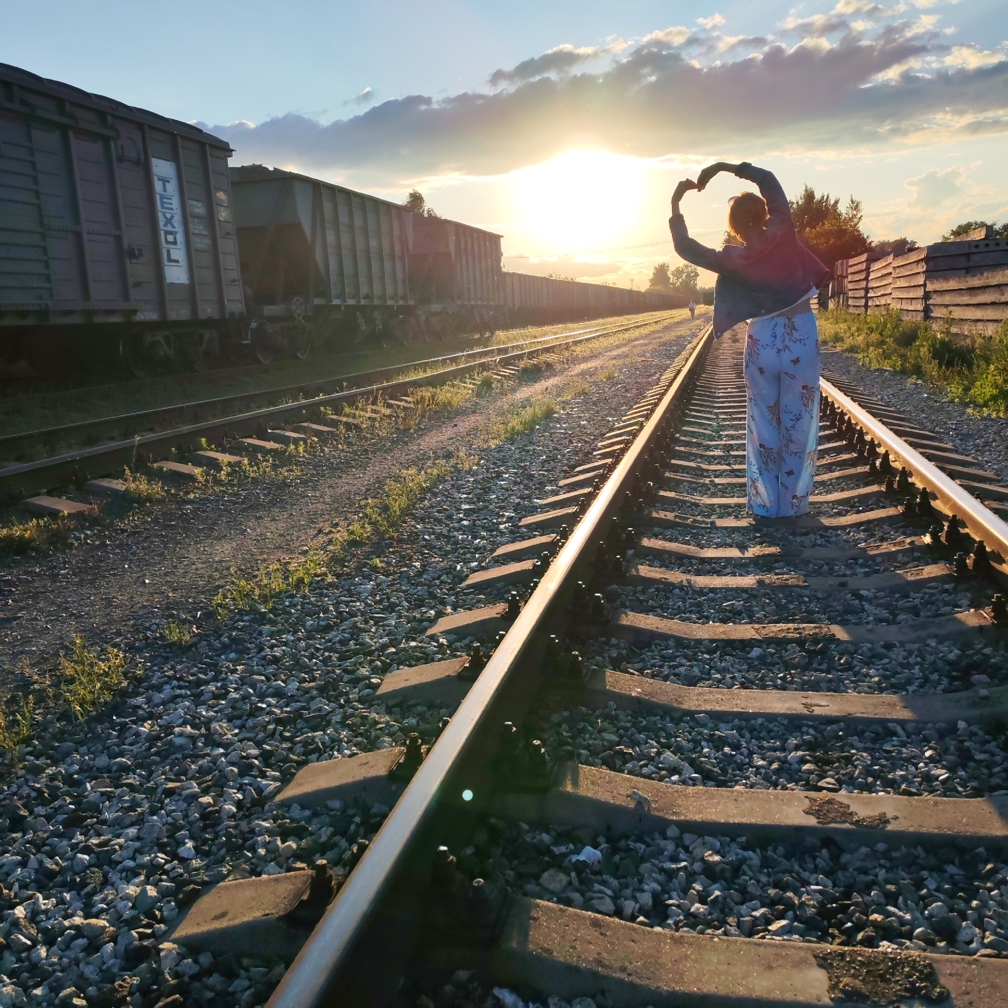 An outdoor, eye-level, wide shot captures a female figure standing on railroad tracks during sunset. The woman is positioned on the right side of the frame, facing away from the camera and towards the setting sun, which is partially obscured by clouds. She is forming a heart shape with her hands above her head. She wears a dark jacket and light-colored pants with a floral pattern. The railroad tracks stretch into the distance, flanked by a long train of dark freight cars on the left and a wooden fence on the right. The sky is a mix of blue and orange hues, with scattered clouds illuminated by the low sun. The ground is covered with gravel and sparse vegetation. A second, smaller figure of a person can be seen further down the tracks in the distance. This image was taken in Chaikovsky, Russia.