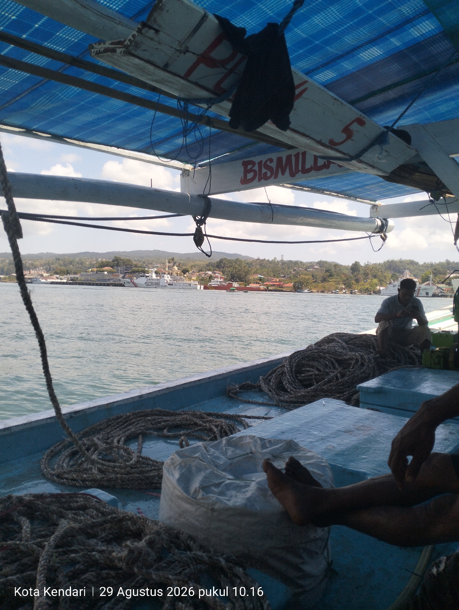 The image depicts a view from a boat, looking towards the coast of Kendari, Indonesia. A man is seated on a pile of ropes on the deck of the boat, holding a mobile phone. The boat has a blue tarpaulin roof supported by wooden beams, with the word "BISMILLAH" painted on one of the beams. In the background, the coastline is visible with buildings and trees, and several ships are docked in the water, including what appears to be a Coast Guard vessel. The water is calm with a slight ripple. The timestamp at the bottom of the image indicates "Kota Kendari | 29 Agustus 2026 pukul 10.16," which translates to "Kendari City | August 29, 2026 at 10:16 AM."