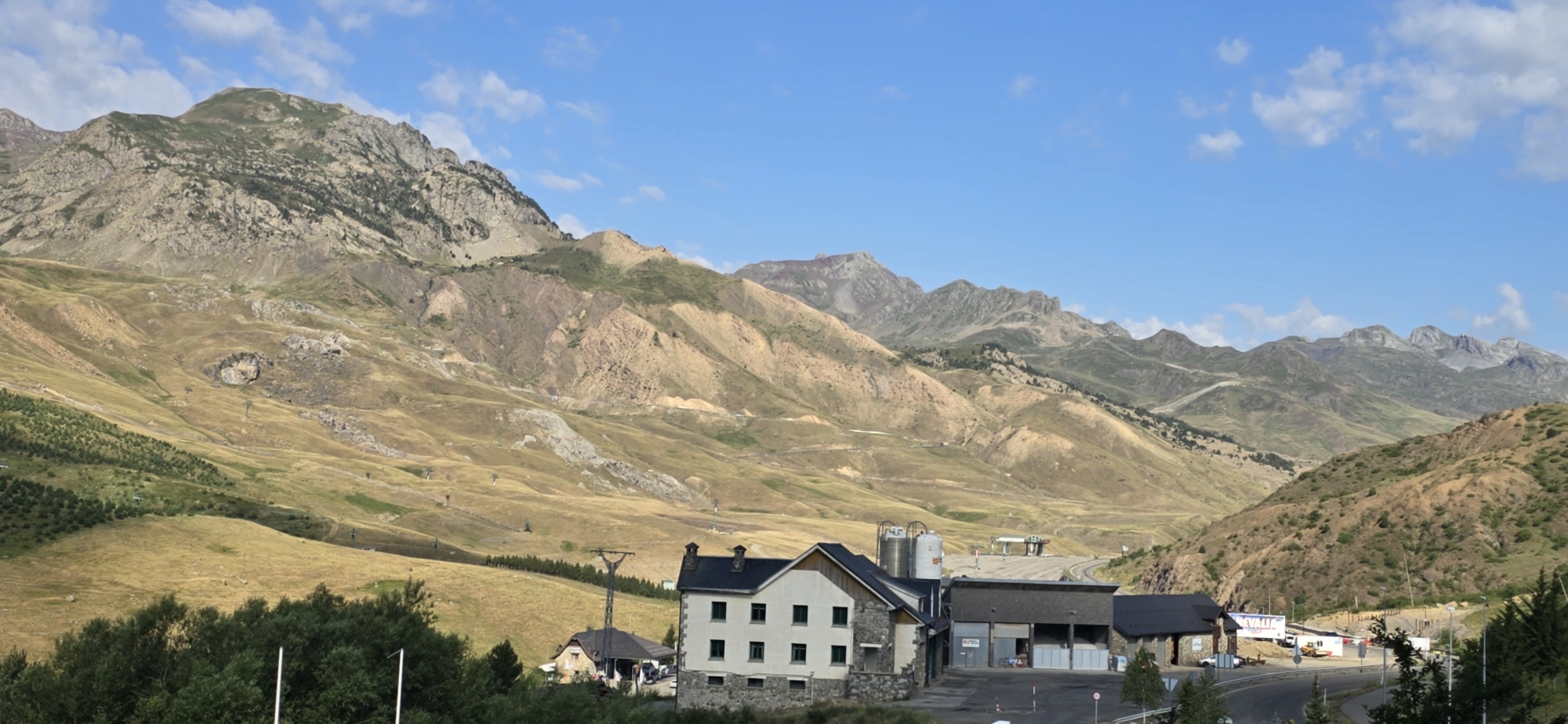 This is a wide-angle, horizontal landscape photograph of a mountainous region under a partly cloudy blue sky. In the foreground, on the left, a dense line of dark green trees partially obscures the view. To the right of the trees, there are several buildings. The most prominent is a white, multi-story building with a dark roof, featuring numerous windows and stone accents at its base. Adjacent to this building, to its right, is a long, dark-roofed structure with open bays. Further right and slightly behind, there are industrial silos and another building. A paved road runs in front of these structures, curving to the right. In the midground and background, rolling hills and steep mountains dominate the scene. The mountains are varied in color, with patches of green grass, dry yellow-brown vegetation, and exposed rock faces showing shades of brown, grey, and ochre. Several ski lift towers are visible on the slopes, suggesting a ski resort area. The overall impression is a semi-arid, mountainous environment with a cluster of buildings situated in a valley. The location is identified as Sallent de Gállego, Spain.